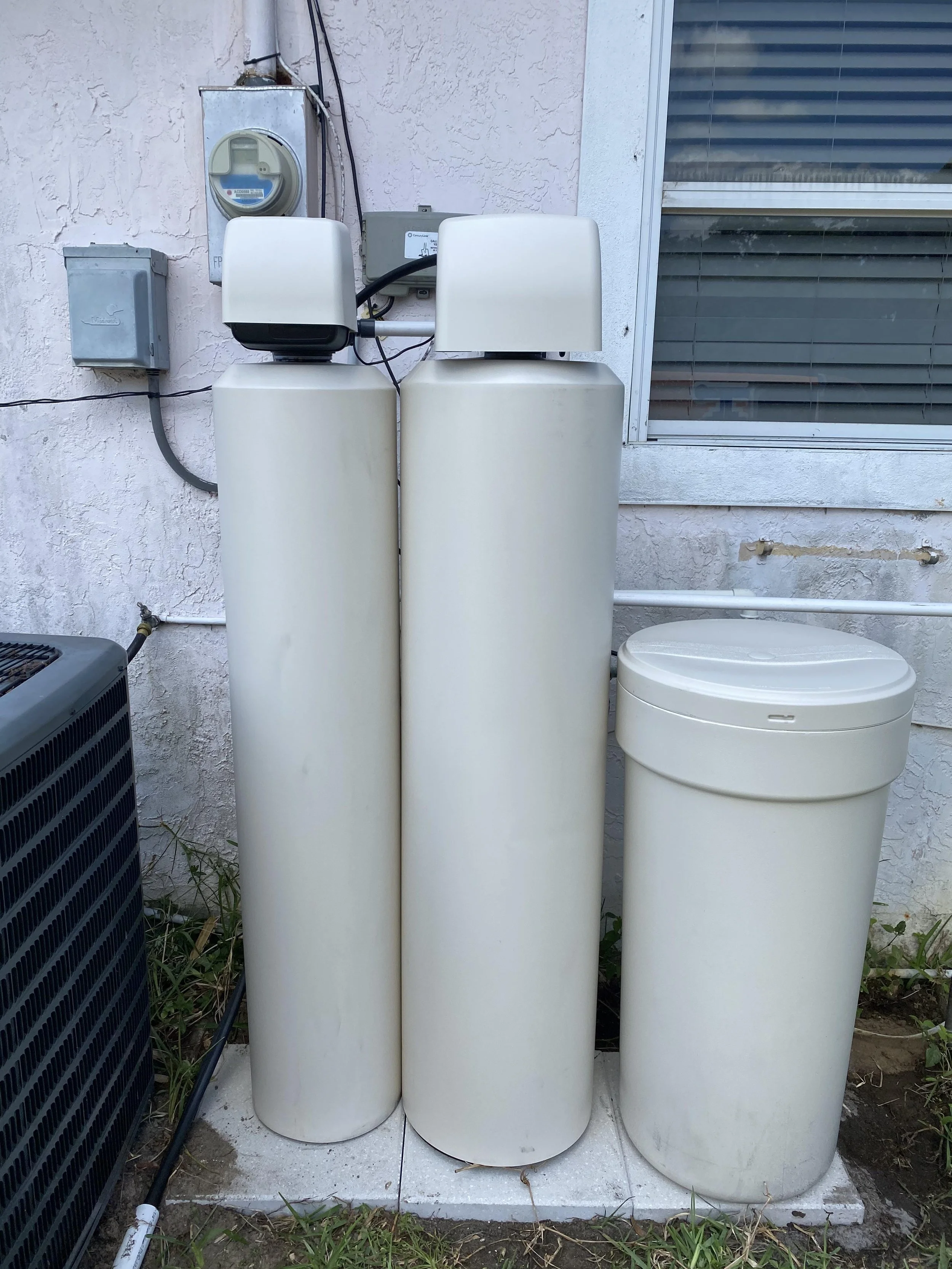 Three large, white water filtration tanks are installed outside a building. To the left, there are electrical boxes and a pipe connected to the tanks. To the right, there is a window with blinds. There is grass and some dirt at the ground level.