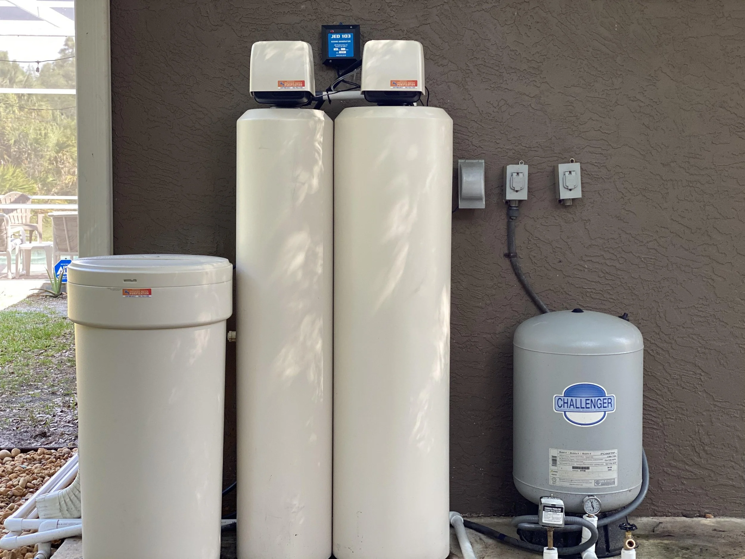 An outdoor water filtration system with five large white cylindrical tanks, a small white round tank in the foreground, and a small white and blue sign labeled 'CHALLENGER'. There are electrical panels and pipes connected to the tanks, against a textured beige wall. In the background, there is a patio with chairs and a table outside a window.