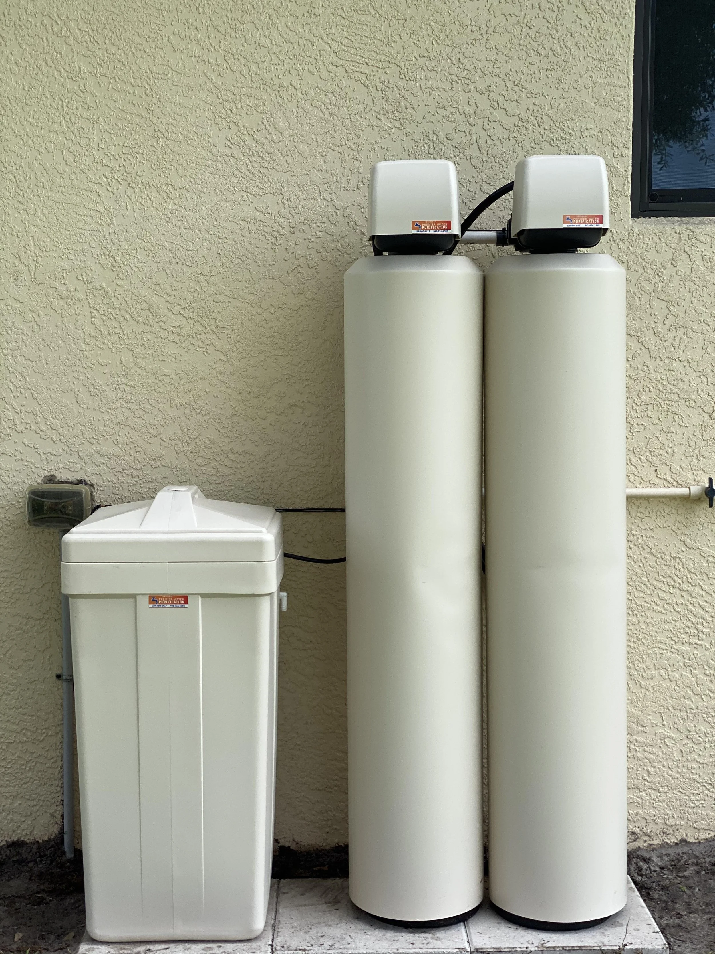 Water filtration system with two large vertical tanks and a salt holding tank on the left, in front of beige textured wall and a nearby window.