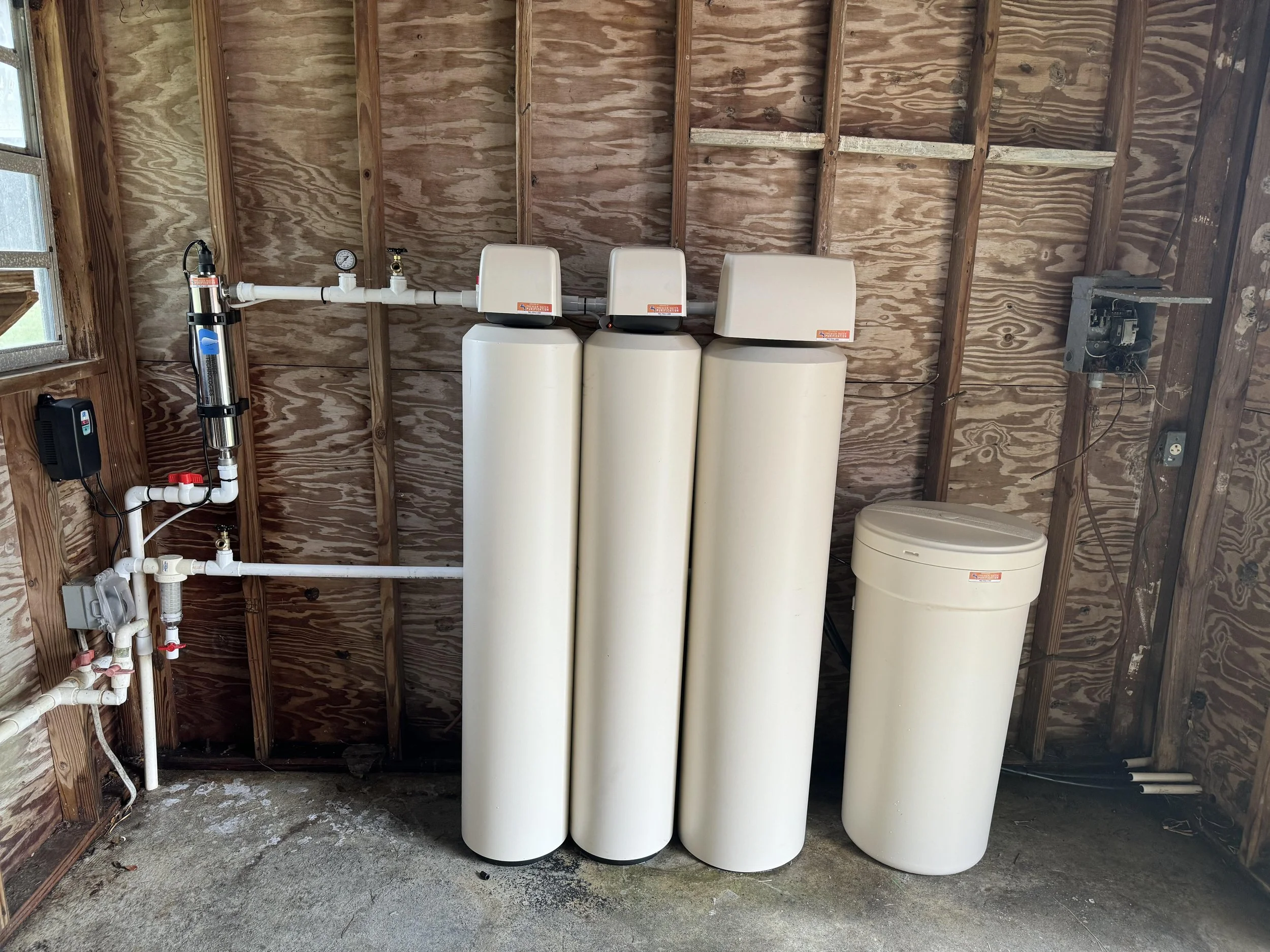 In an outdoor shed is three tanks for water filtration, with a salt tank to the far right. to the left is an UV light for extra water purification.