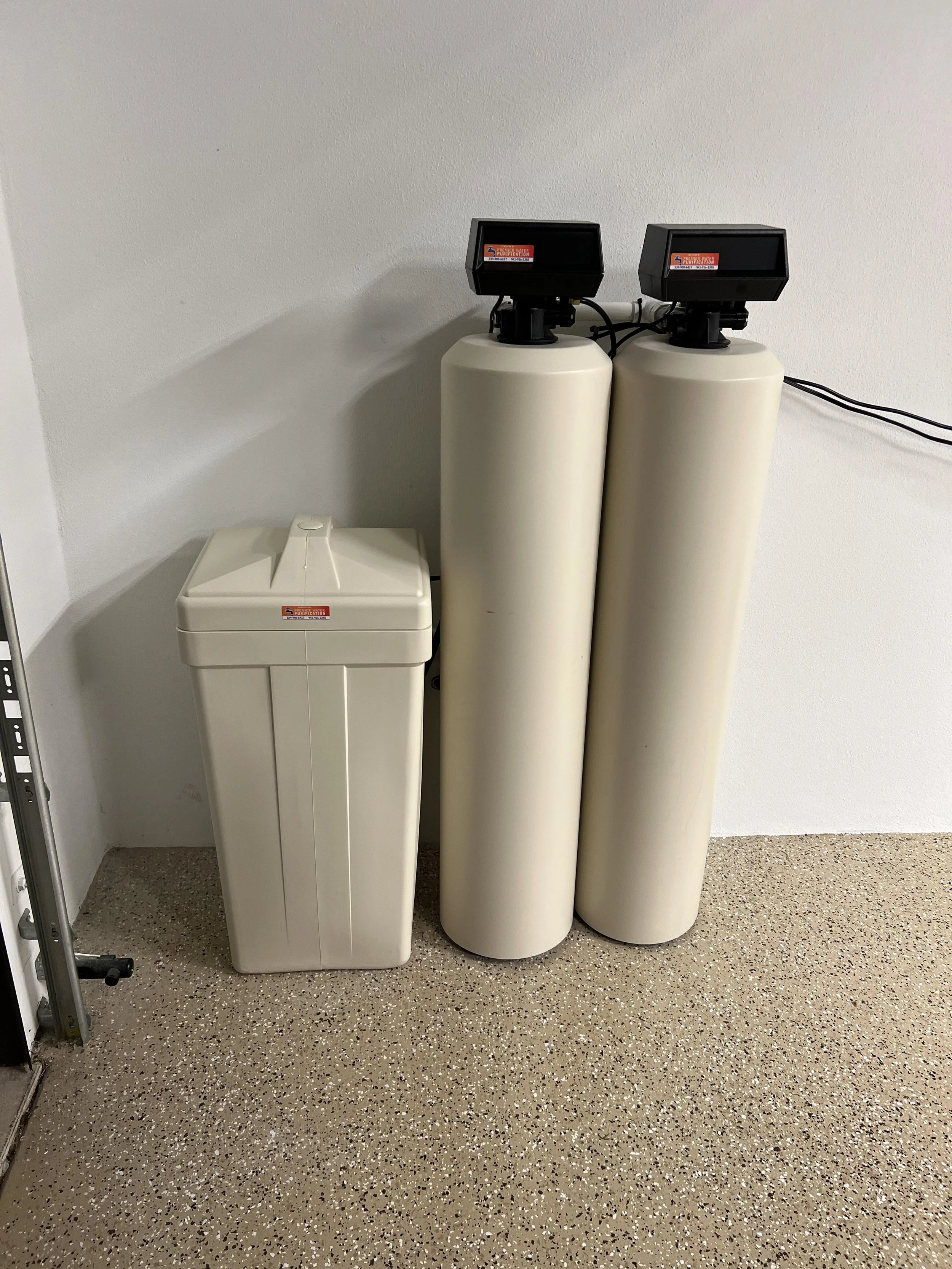 Water filtration system with three large beige tanks against a plain wall and speckled brown floor.