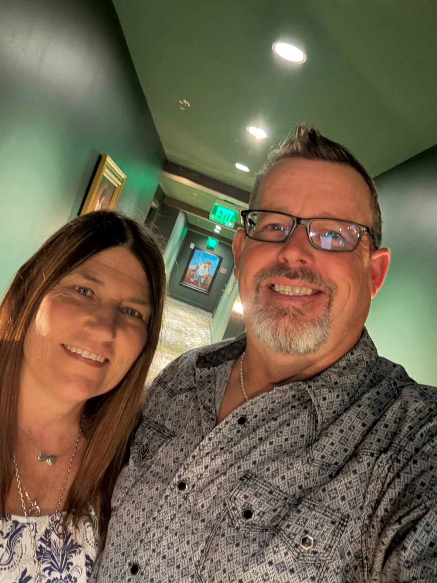 Close-up of a smiling woman with long brown hair and a man with glasses, a gray beard, and a patterned shirt, taking a selfie in an indoor setting with green walls, an exit sign, and a painting in the background.