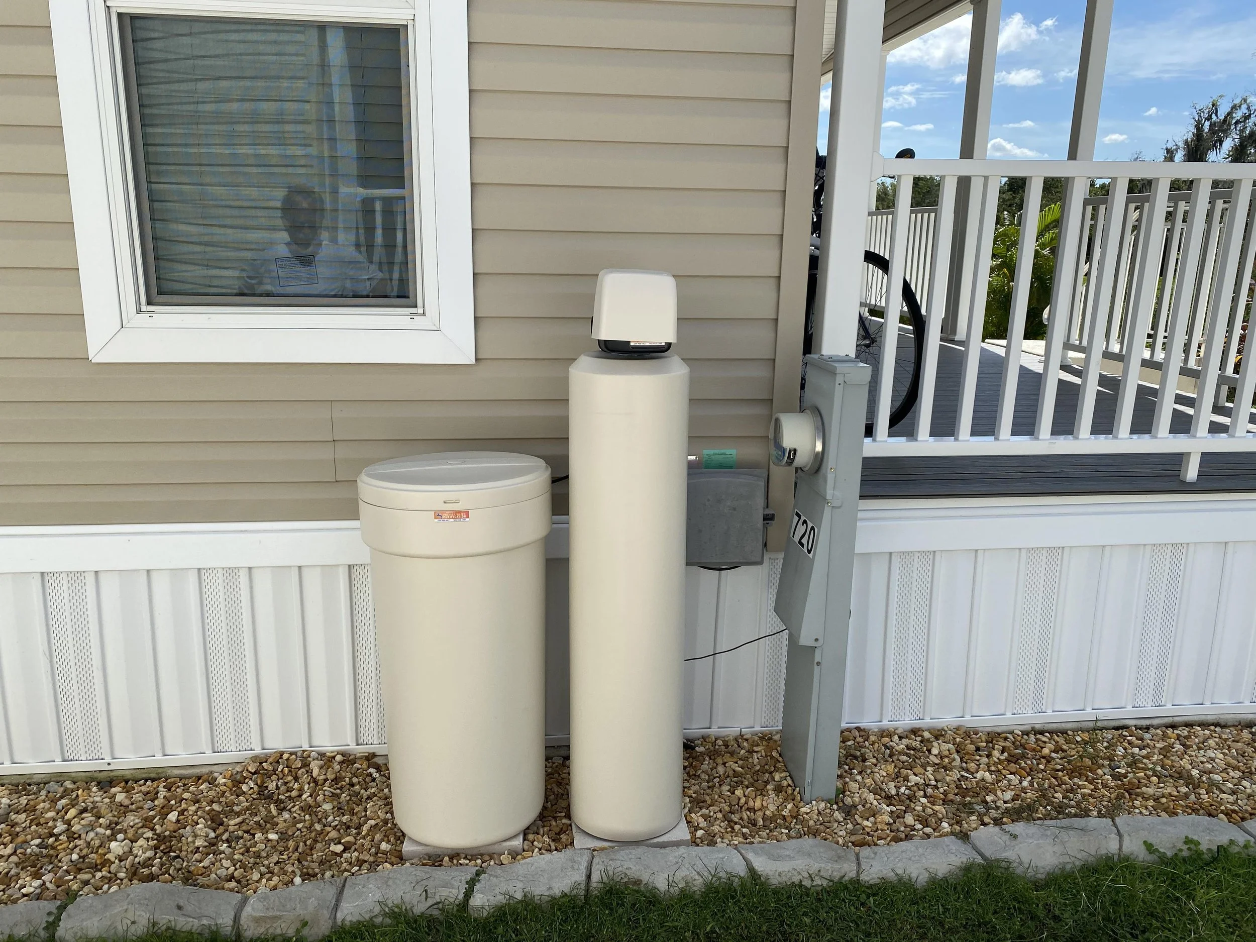 The image shows the outside of a house with beige vinyl siding. There is a window on the left. There is one beige water filtration tank with a salt container to the left of it. 