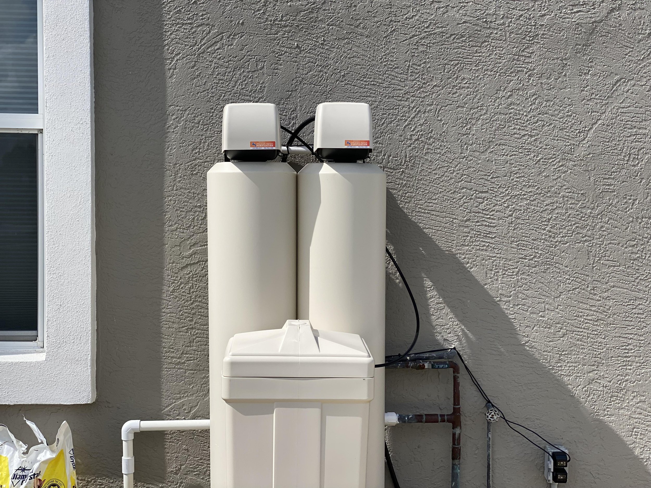 Water filtration system installed on an exterior wall next to a window, with pipes and electrical connections at the bottom.