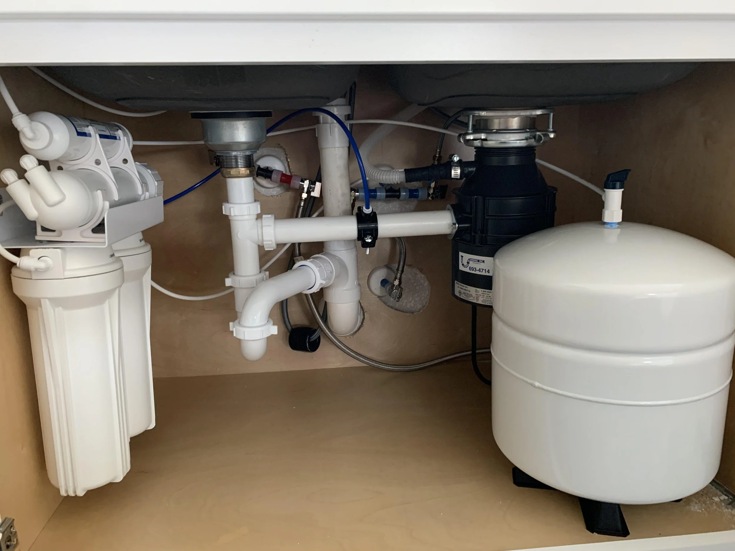 Under-sink plumbing including filters, pipes, in a kitchen cabinet.