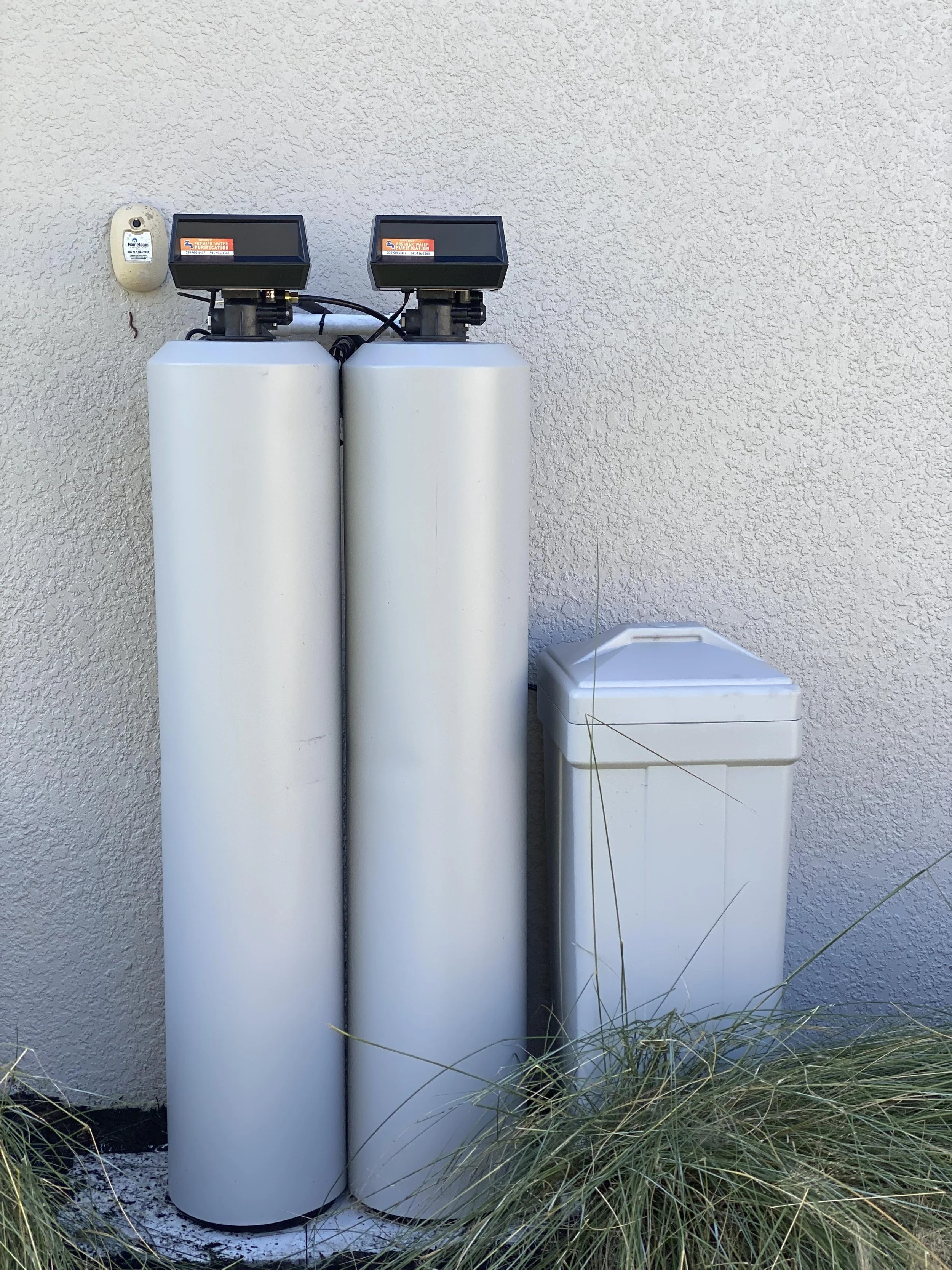 Two white water softening tanks with digital control units on top, mounted on a wall outside next to a beige wall, surrounded by green grass.