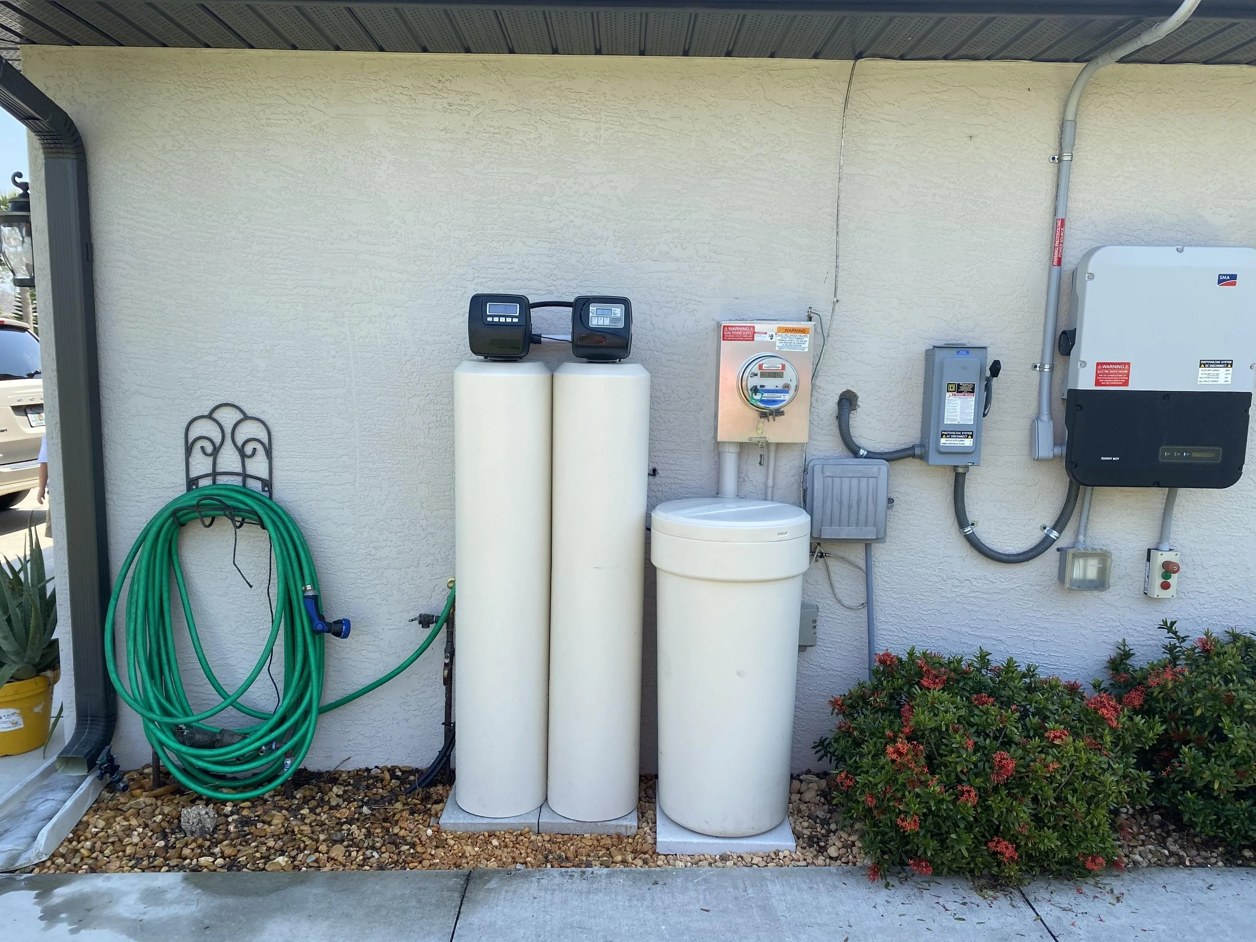 A utility area on the side of a house with water treatment equipment, including a water softener, a meter, and other electrical components mounted on the wall. There are green garden hoses hanging, a yellow flower pot, and red flowering bushes in the