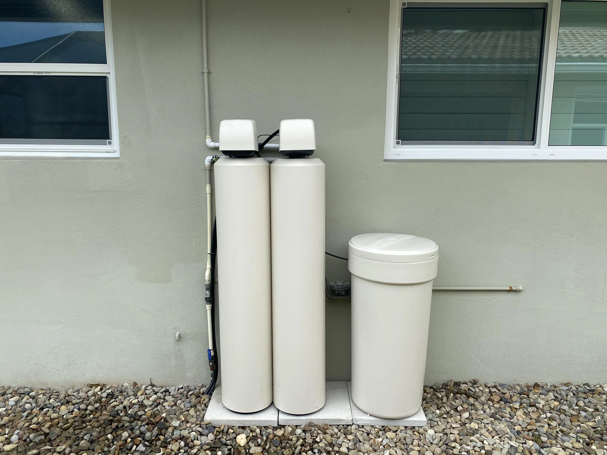 Outside view of water treatment or filtration system with three large tan tanks, positioned against a light green wall between two windows, surrounded by a bed of small rocks.