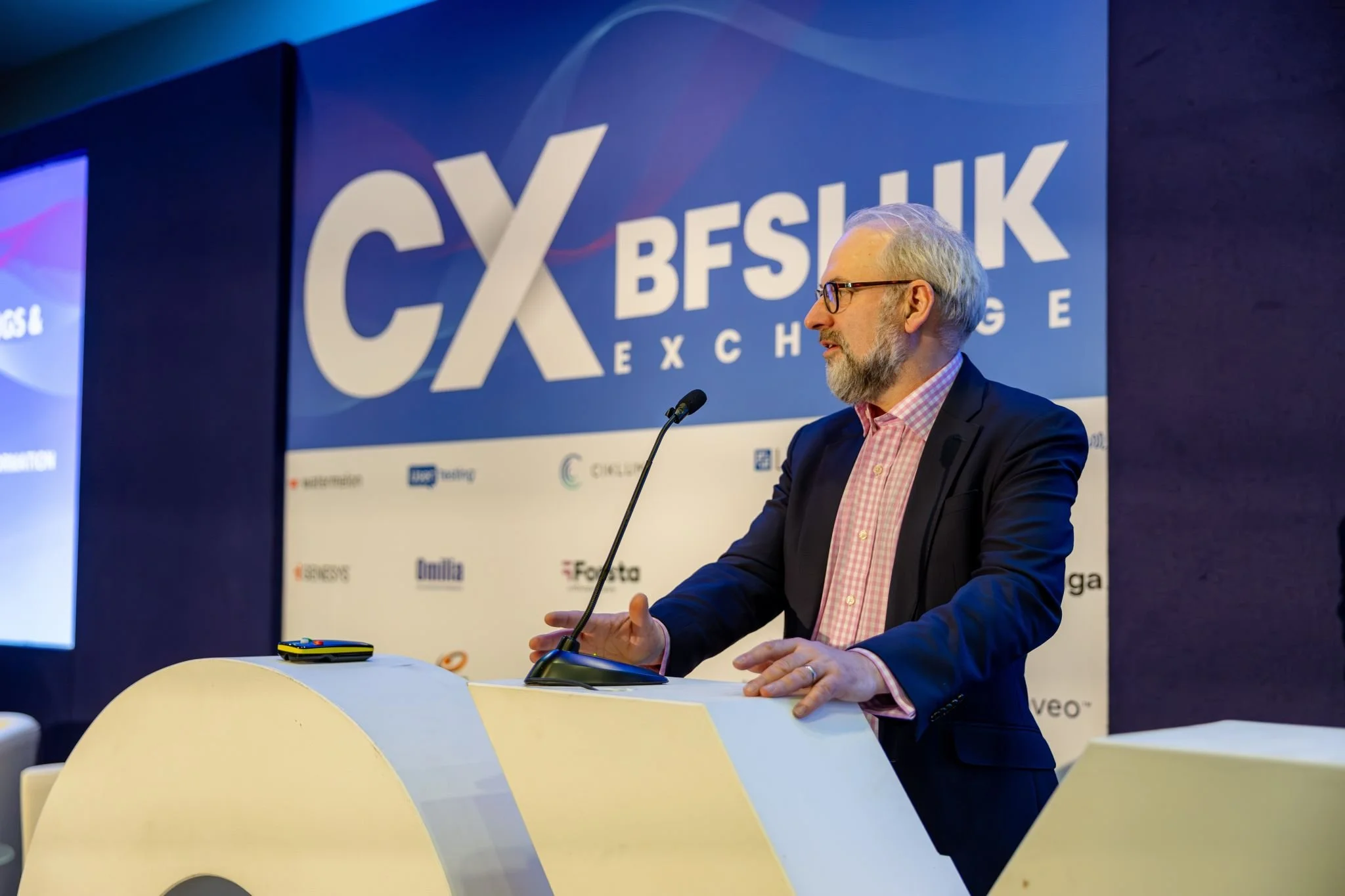 A man with a beard and glasses wearing a pink checkered shirt and dark blazer speaking at a conference or event. He stands at a podium in front of a large blue banner with the text 'CXL BESUIT' and other smaller sponsor logos.