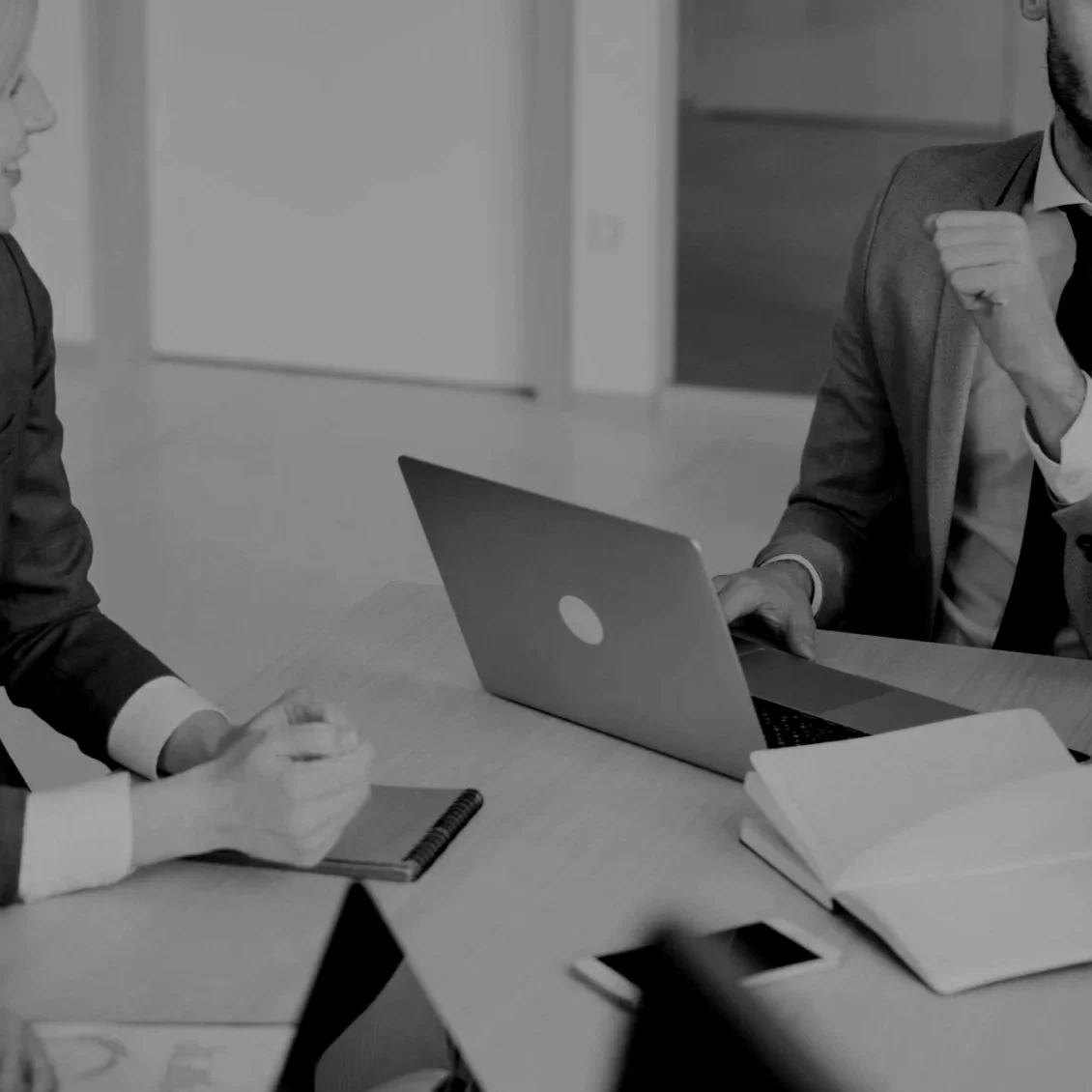 People in business attire engaged in a discussion at a meeting table, with a laptop, notebooks, papers, and a smartphone on the table.