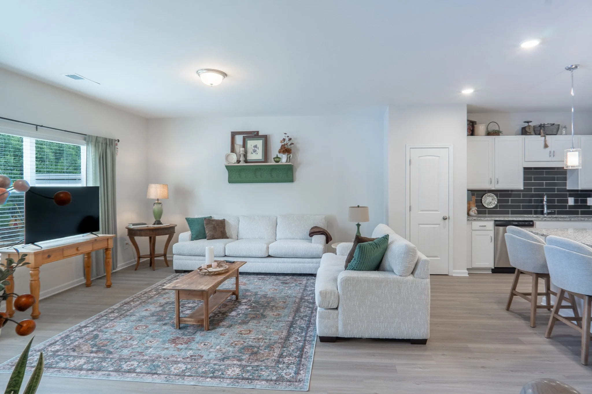 A cozy living room with white walls and a large window with blinds and curtains. There's a white sofa with cushions, a wooden coffee table on a patterned rug, and a living room lamp. The adjacent kitchen has white cabinets, a black backsplash, and a 