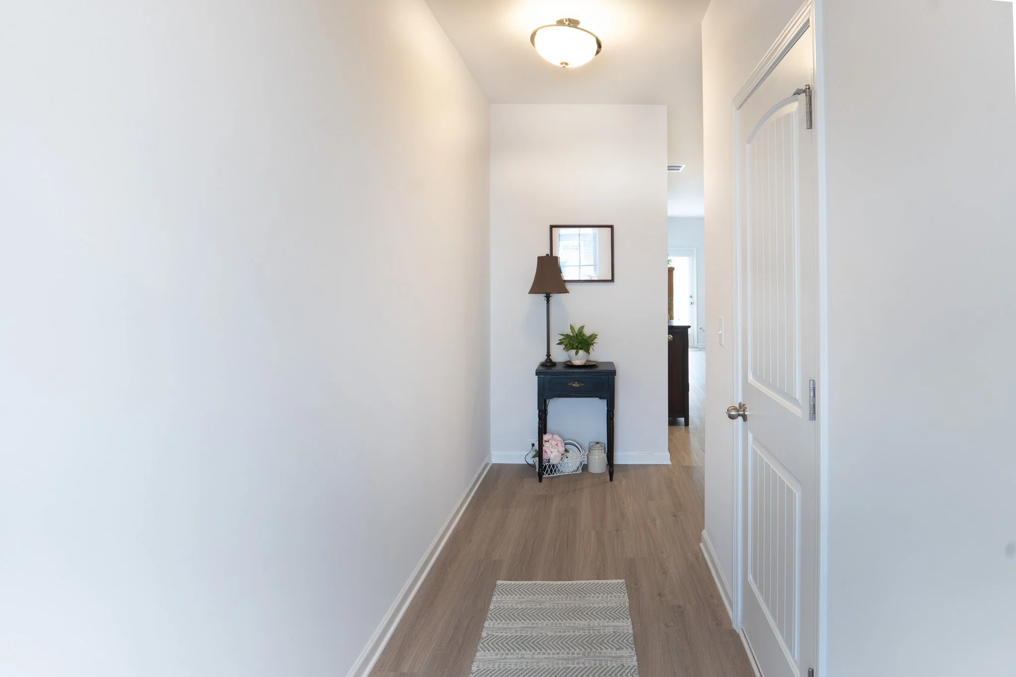 Entryway with beige walls, a small black table with a lamp, mirror, plant, and decor items, with a hallway leading to other rooms.