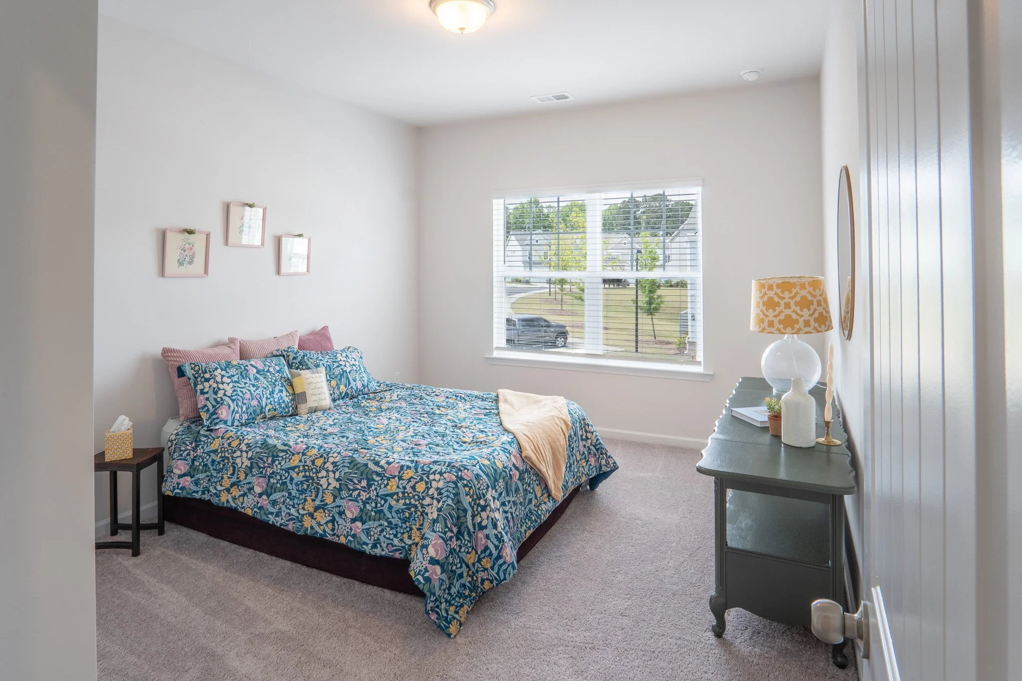 A cozy bedroom with a bed covered in a blue floral quilt, pink and blue pillows, white walls, a window with blinds showing a suburban neighborhood, a gray dresser with a lamp and decorative items, and a small nightstand with tissue.