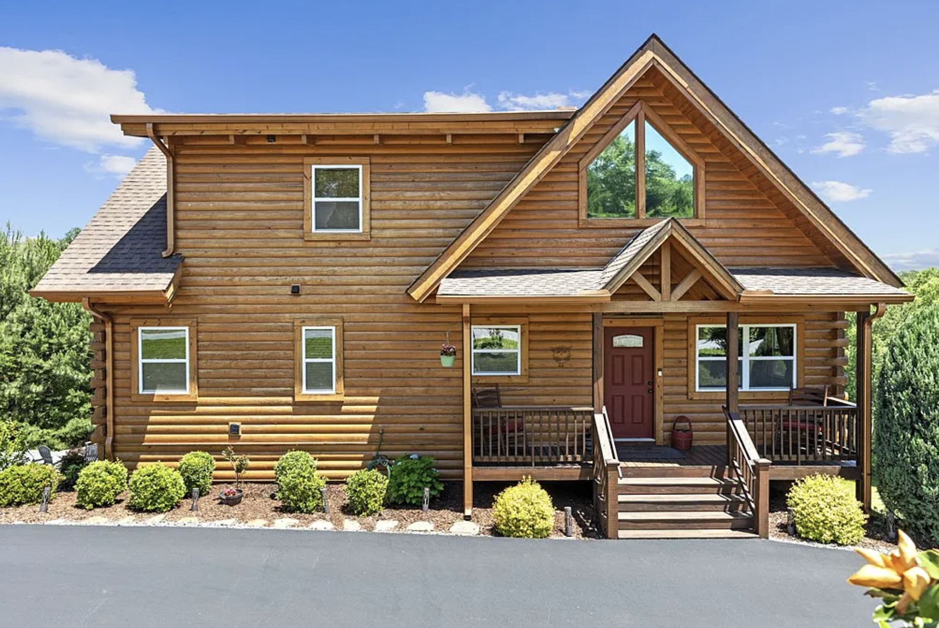 Front view of a two-story wooden house with a red door, screened porch, multiple windows, and a triangular gable with large window, surrounded by green bushes and trees, under a blue sky with clouds.