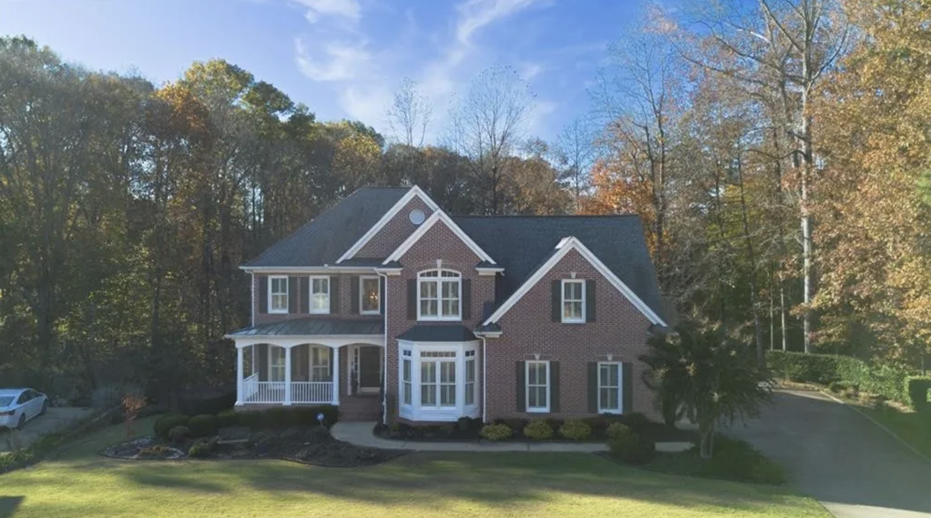 A two-story brick house with a white front porch and multiple large windows, surrounded by well-maintained lawns and trees with autumn foliage, under a partly cloudy sky.