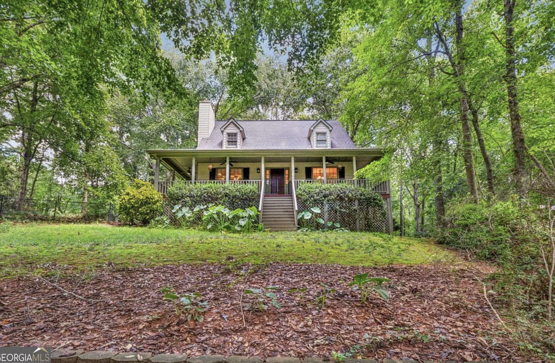 A two-story house with a large porch, surrounded by tall green trees. The house has a steep gabled roof with two small dormer windows, black shutters, and brick chimneys. A staircase leads up to the porch from a grassy yard with mulch and small plants.