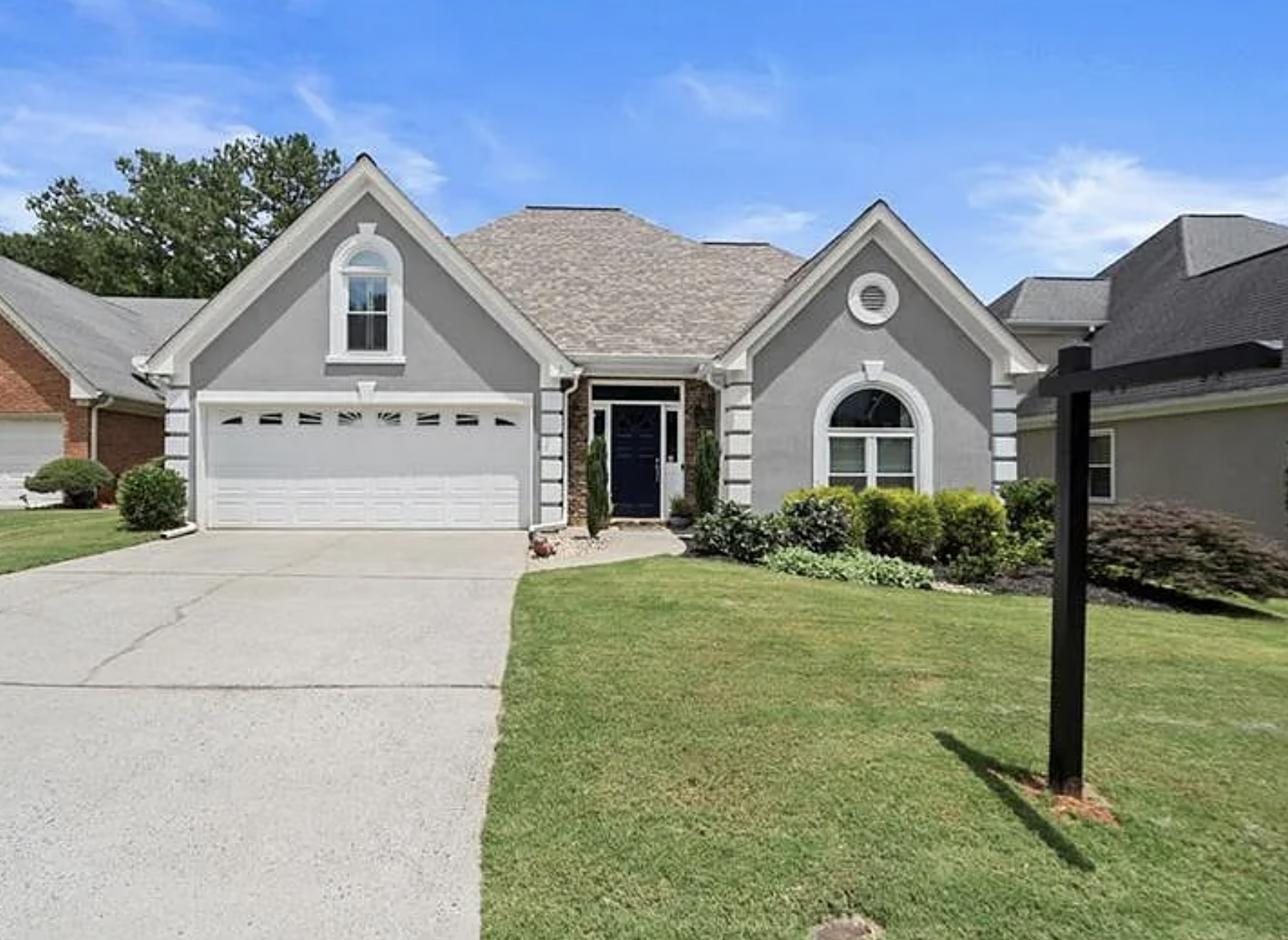 Front view of a gray house with a white garage door, manicured lawn, and a landscape with bushes and trees under a blue sky.