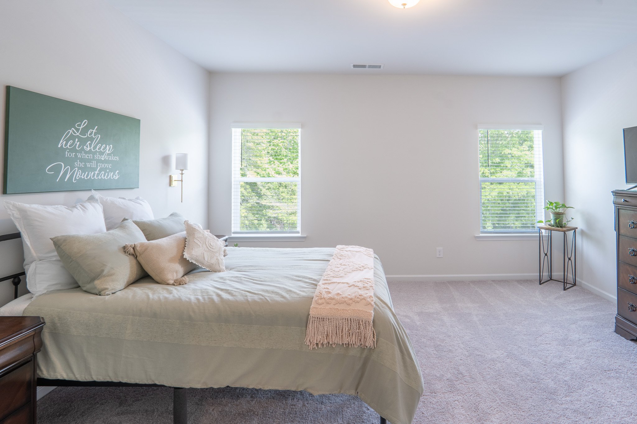 Bright bedroom with a large bed, white bedding, beige pillows, blush throw, two windows with blinds showing green trees outside, a wall-mounted lamp, a framed quote, a small side table with a plant, and a wooden dresser with a TV.