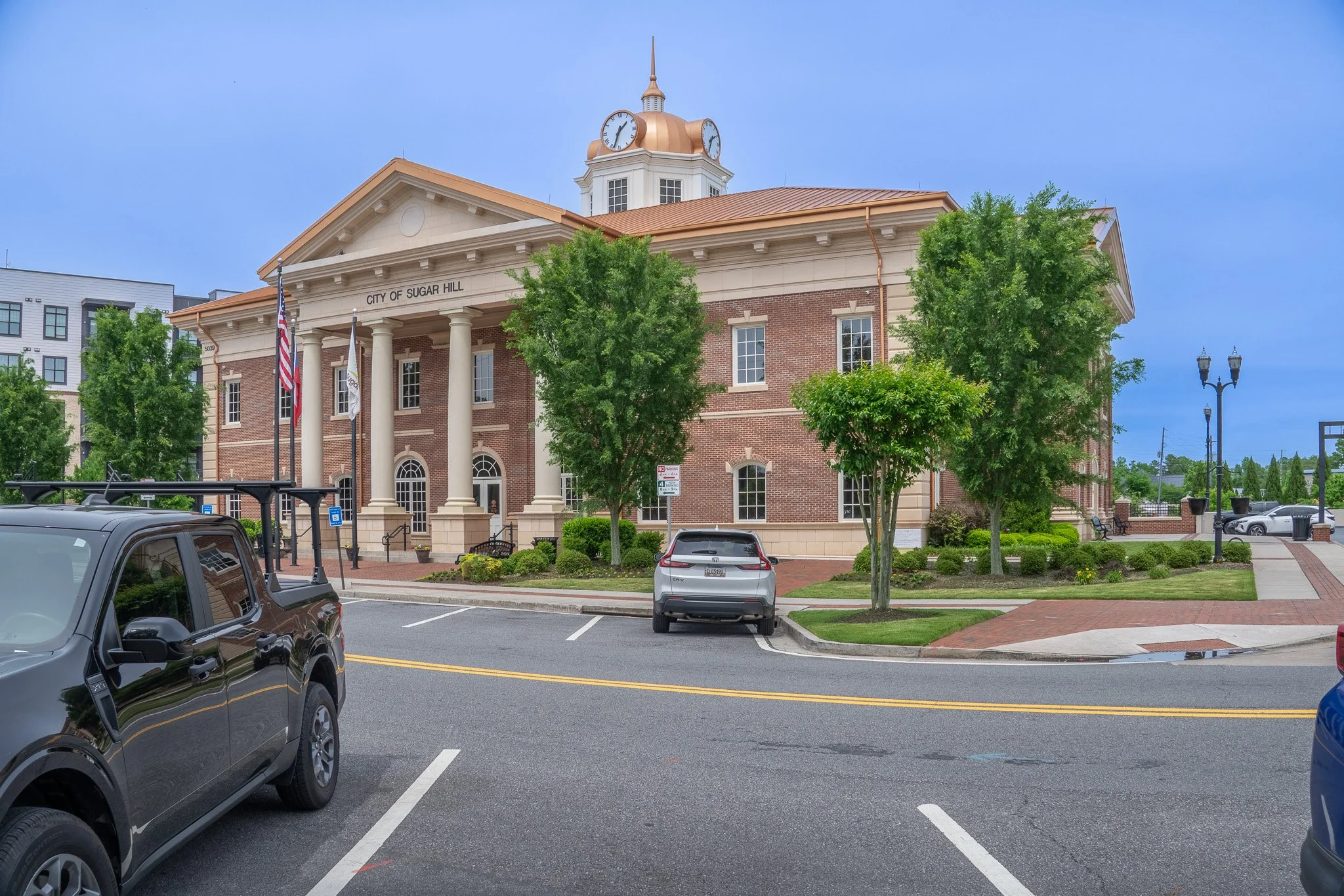 The image shows a historic brick building with a clock tower, labeled 'City of Sugar Hill.' It has white columns at the entrance, flags, trees, and parked cars in the foreground.