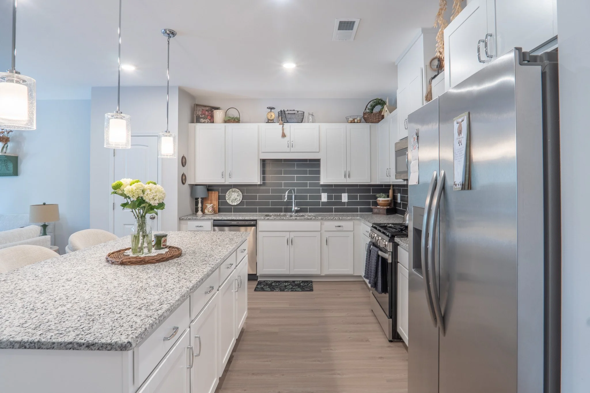 A modern kitchen with white cabinets, gray tile backsplash, stainless steel appliances, and a speckled granite island. Decor includes a vase of white flowers and hanging pendant lights.