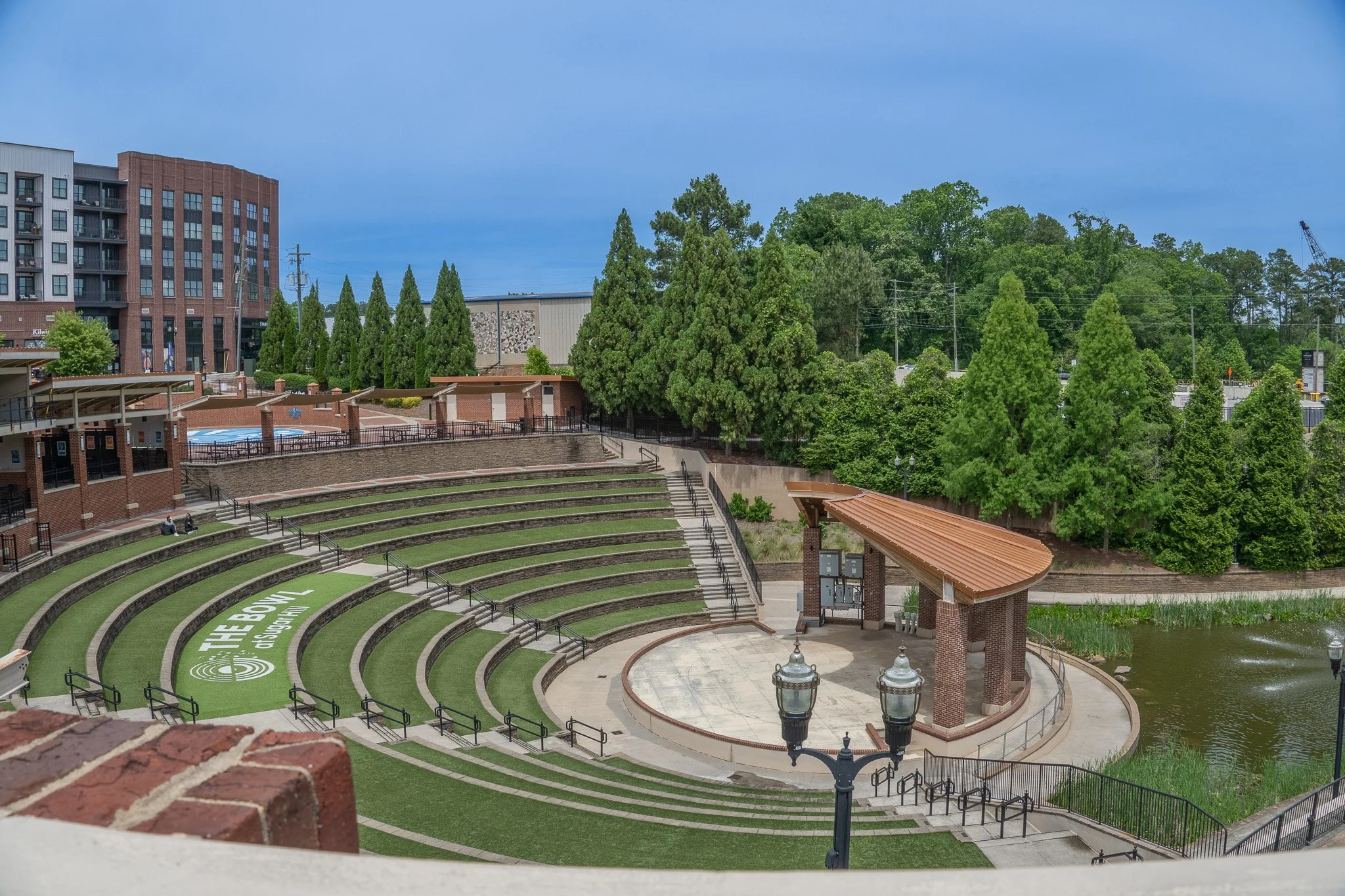 An outdoor amphitheater with ascending green tiers and a paved stage area, surrounded by trees and nearby buildings, with a clear blue sky overhead.