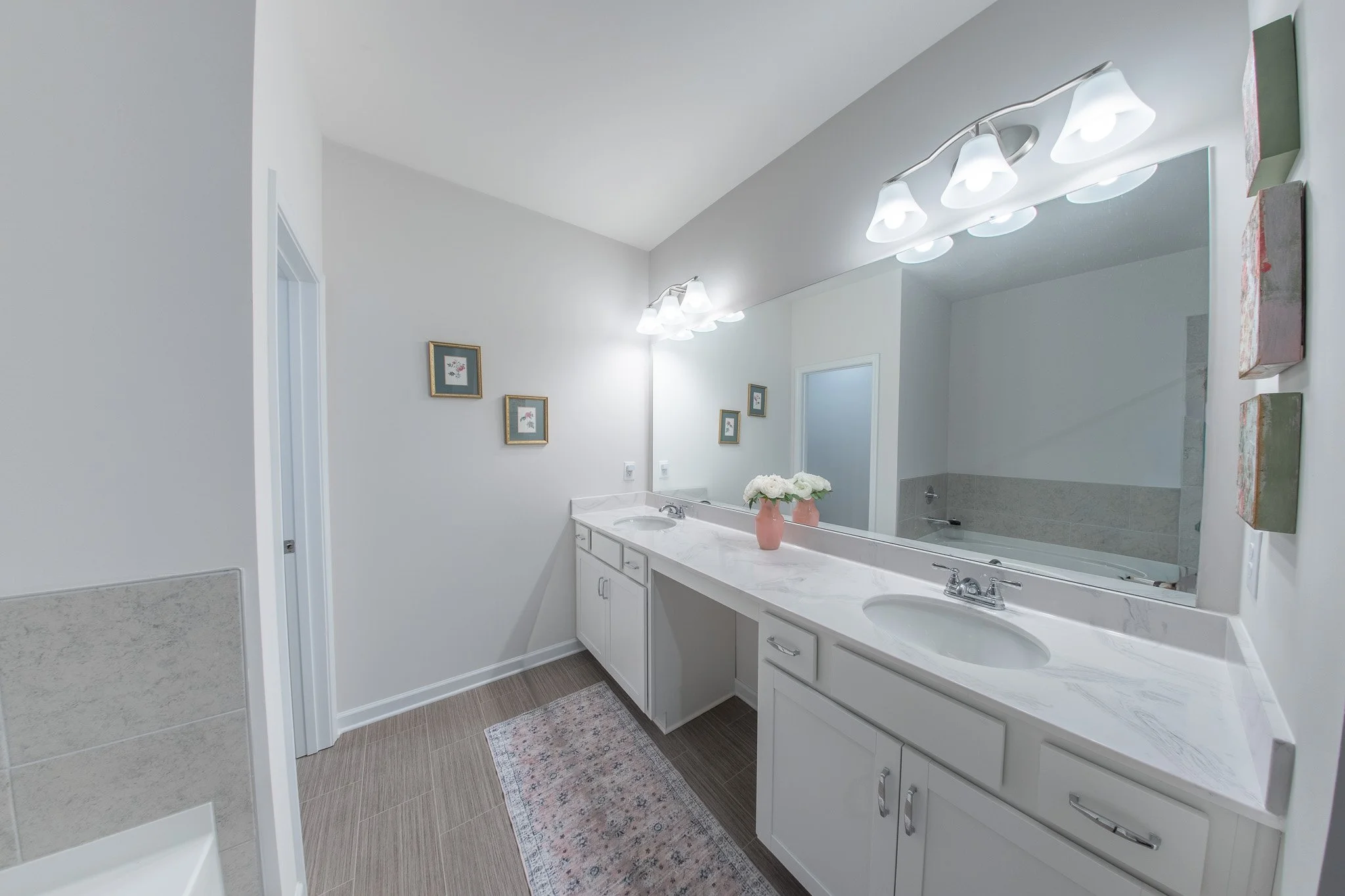 A bathroom with a large mirror above a double sink vanity, white cabinets, framed artwork on the wall, a small rug, and decorative vases on the countertop. The lighting fixtures have five white lamps.