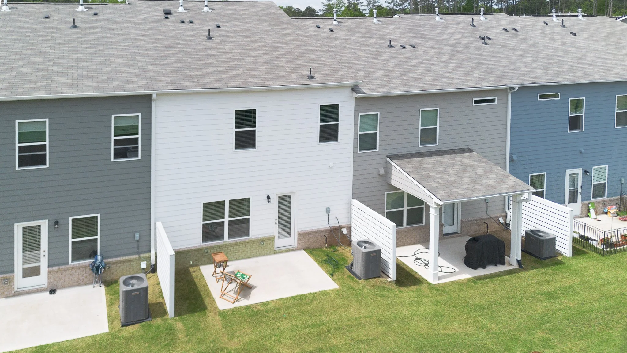 Aerial view of a row of modern townhouses with gray, white, and blue siding, multiple windows, backyards, and air conditioning units on concrete slabs.