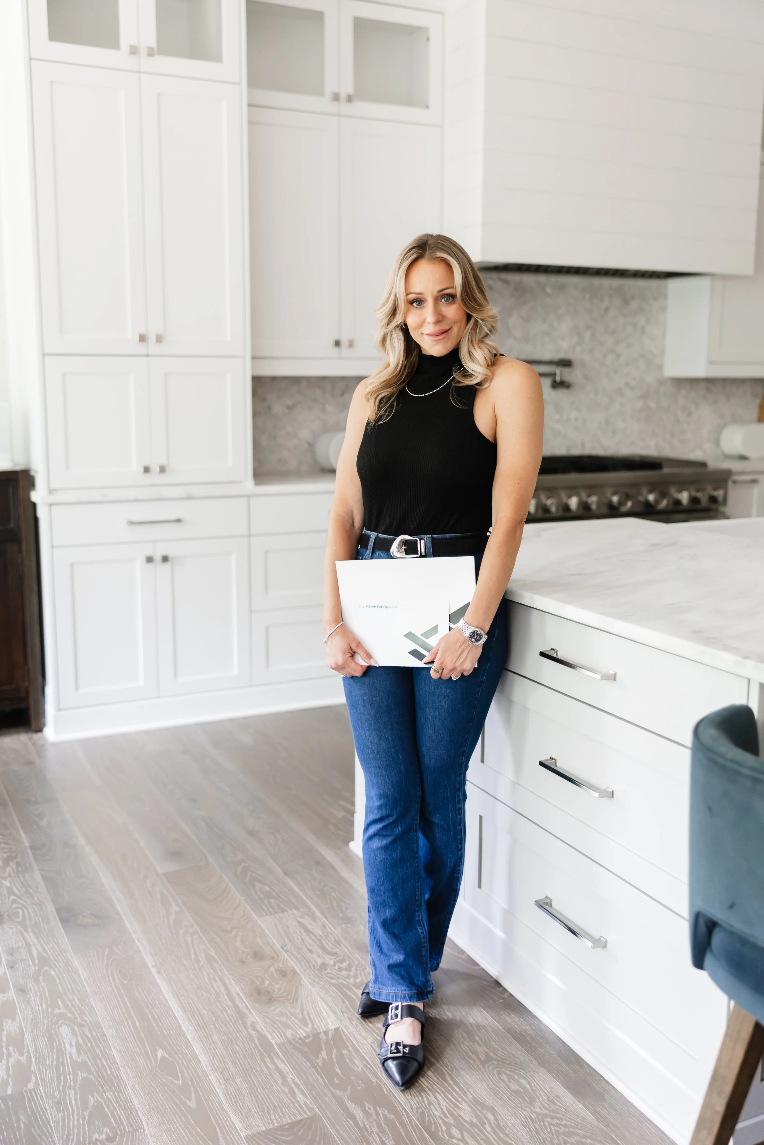 A woman with blonde hair wearing a black sleeveless turtleneck, blue jeans, black shoes with buckles, and a watch, holding a document or magazine, standing in a modern white kitchen with cabinets, a stove, and light-colored wooden flooring.