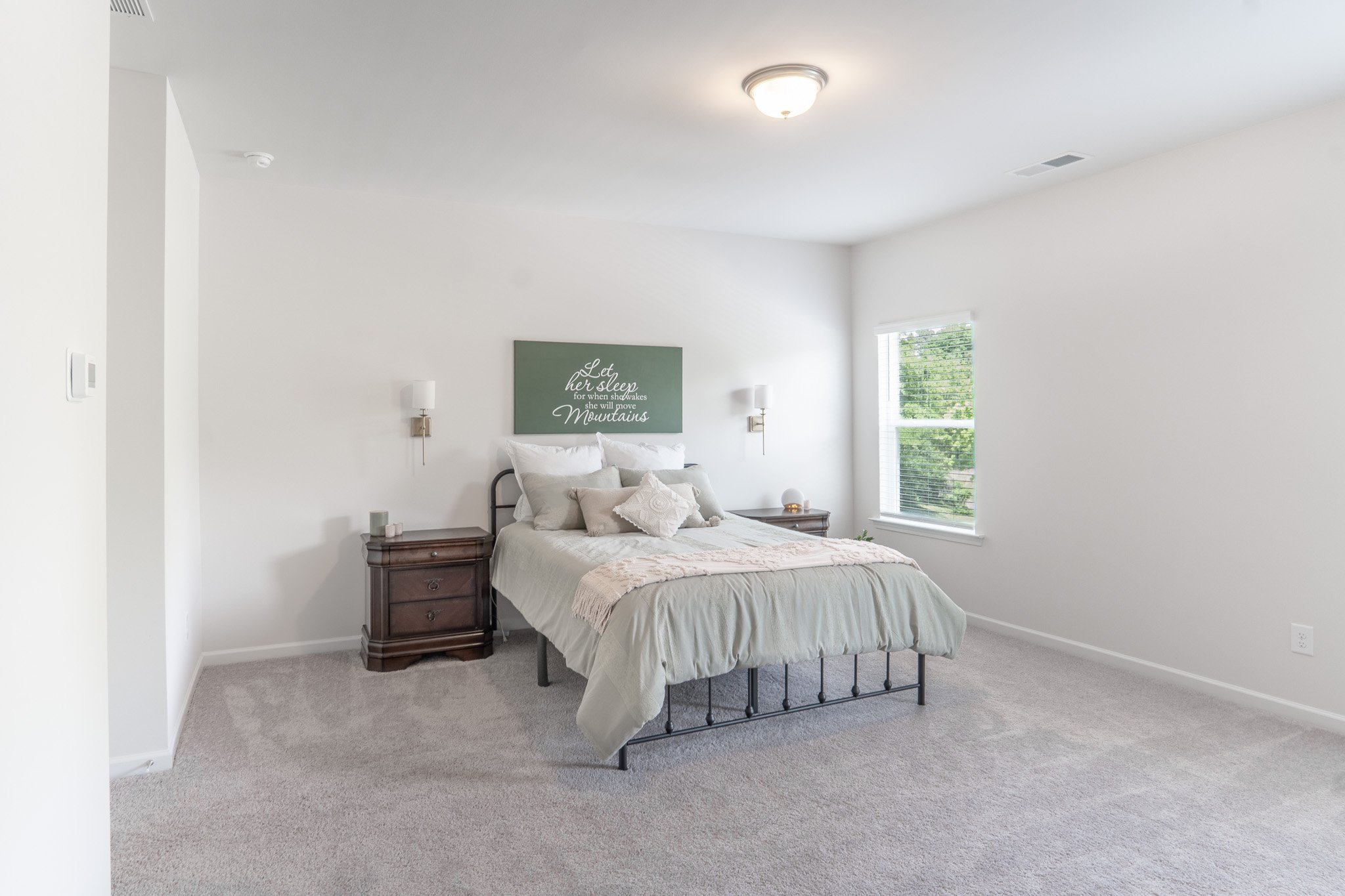 A bedroom with a bed, two nightstands, and a window, decorated with a sign that reads 'Let her sleep for when she wakes, she will move mountains'.