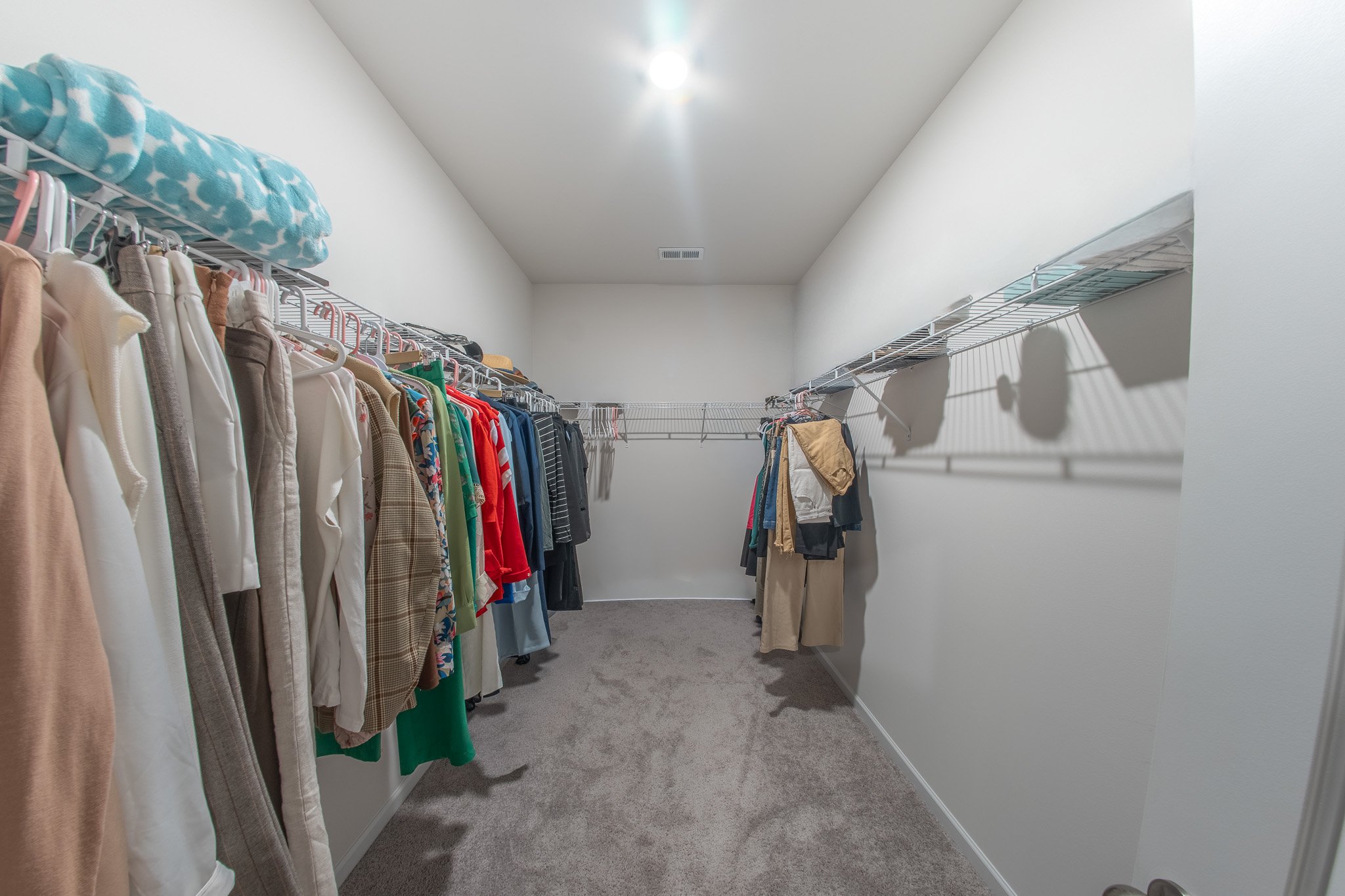 Empty walk-in closet with clothing hanging on racks on both sides, white walls, gray carpet, and a ceiling light.