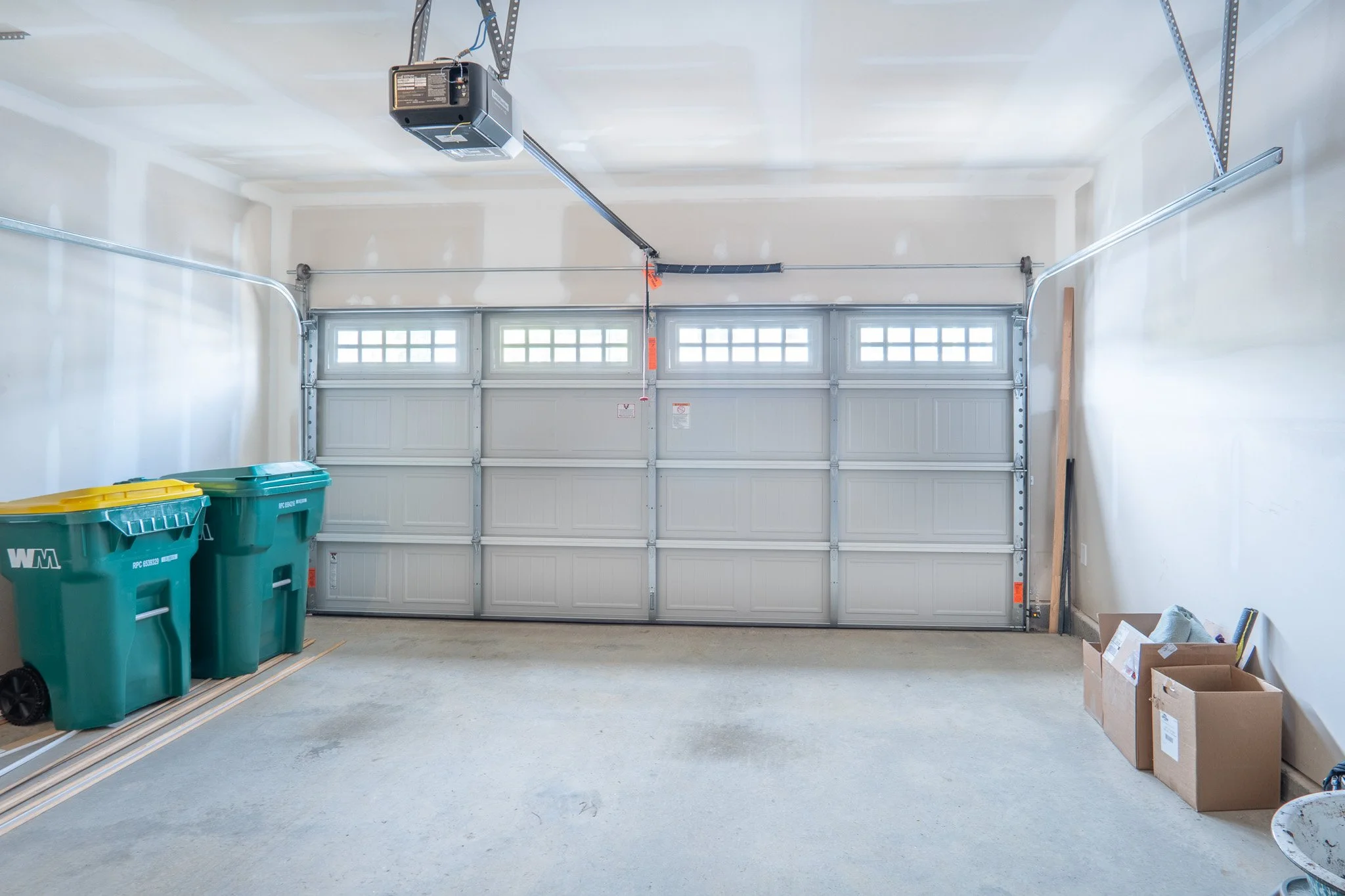 An empty garage with a closed garage door, two green recycling bins, a cardboard box, and a wooden plank against the wall.