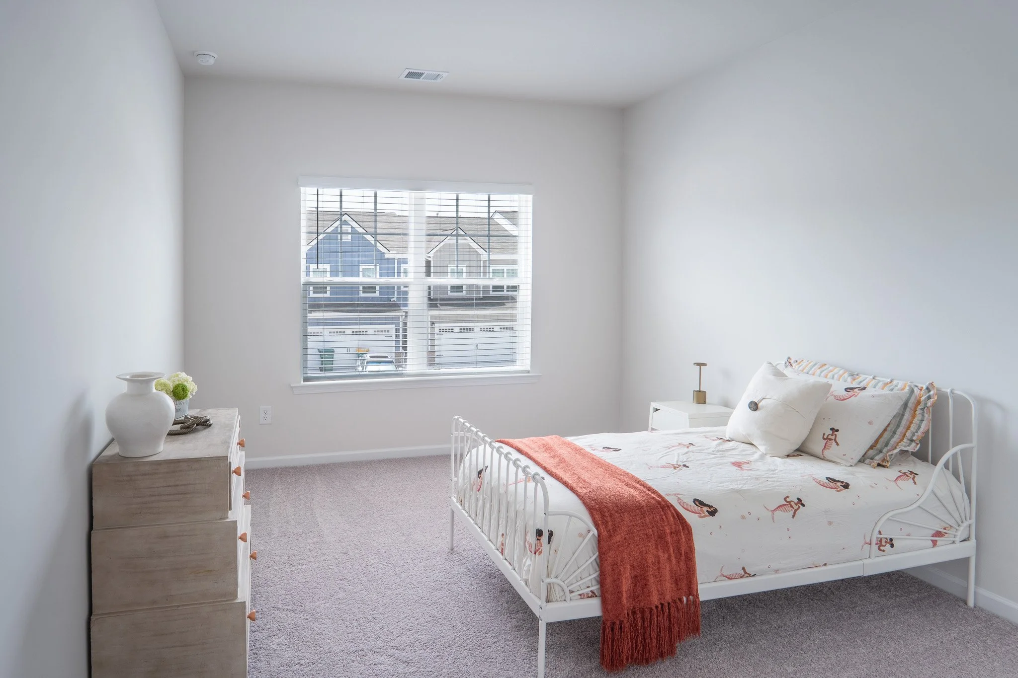 A bright, neatly arranged bedroom with white walls, a window with blinds showing neighboring houses, a white bed with a flamingo-themed quilt, a red throw blanket, and pillows, a wooden dresser with a vase and flowers, and a nightstand with a small l