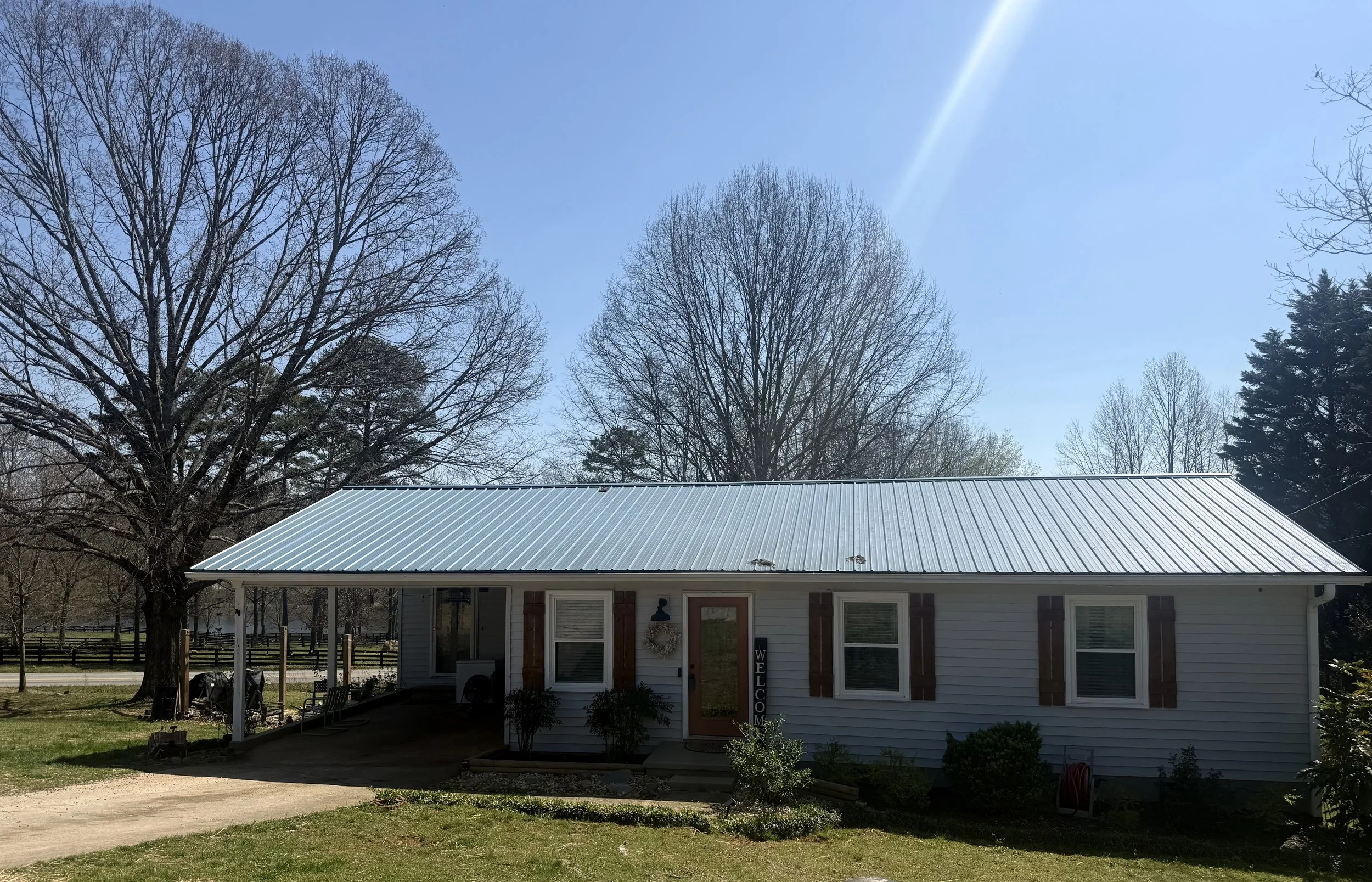 A house with a metal roof, surrounded by trees and a small garden, under a clear blue sky with the sun shining.