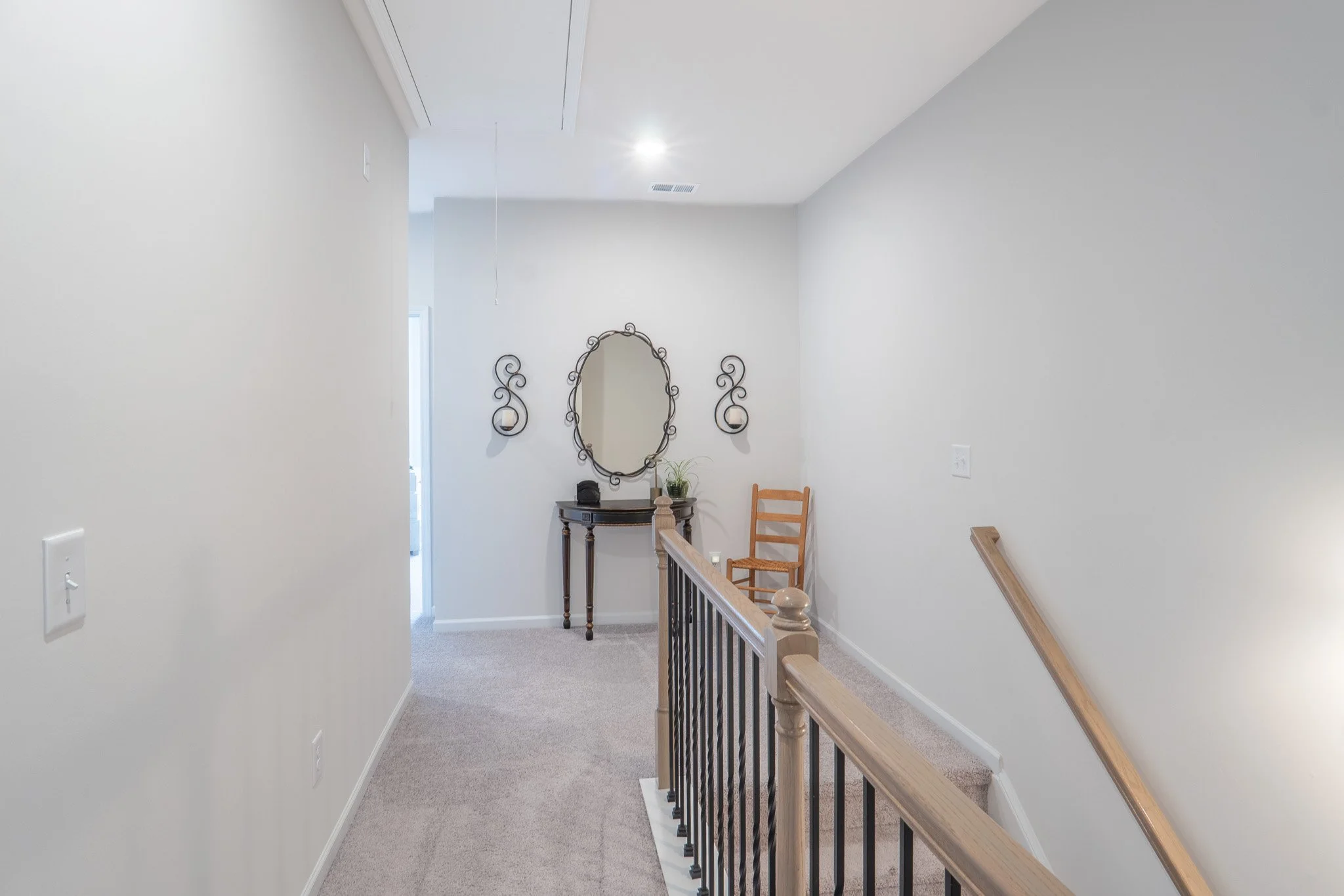 A hallway with off-white walls, beige carpet, a wooden staircase with black metal balusters, a decorative mirror with two candle holders on either side, a small table with a plant and item on top, and a chair. Ceiling has a white air vent and a hangi