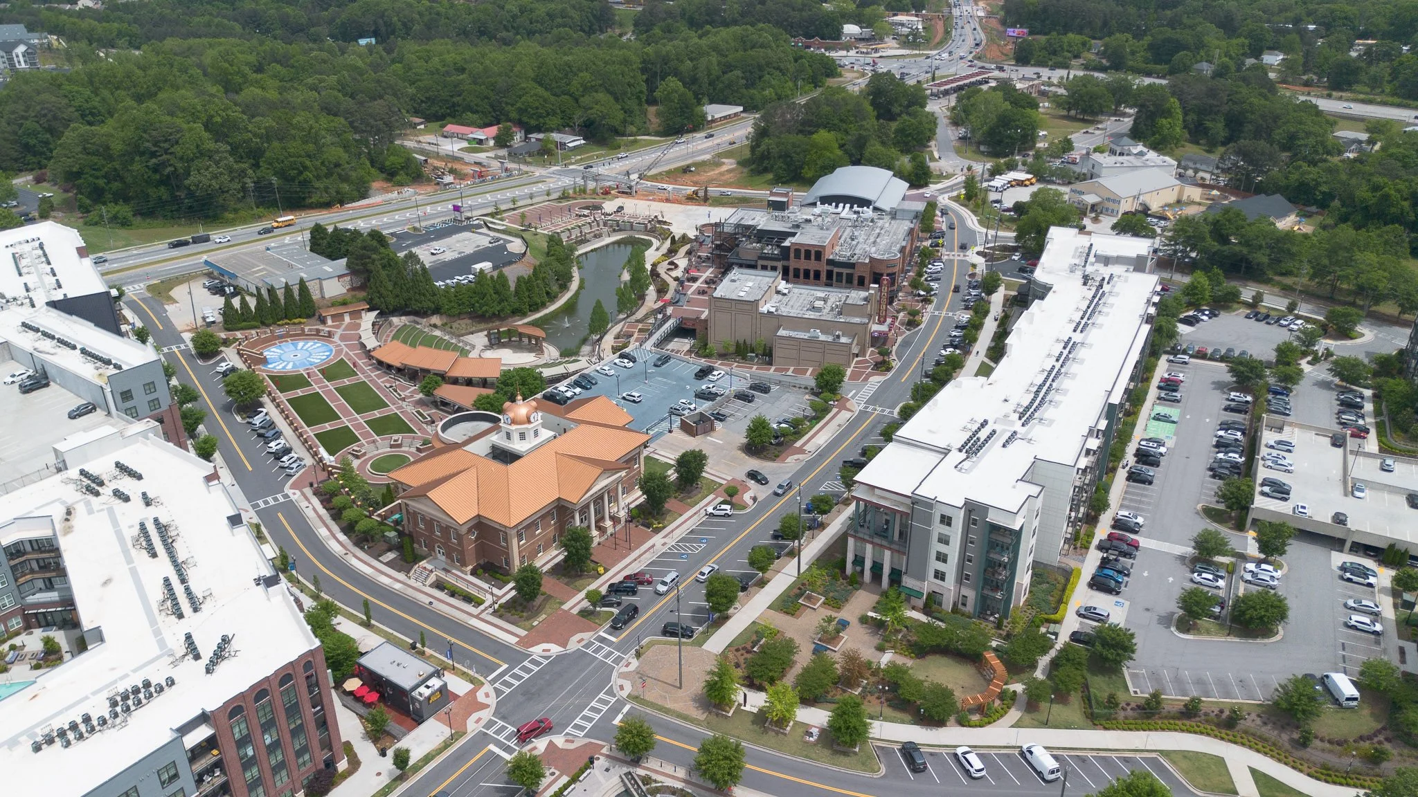 Aerial view of a mixed-use development with roads, parking lots, a water feature, greenery, and buildings in an urban area.