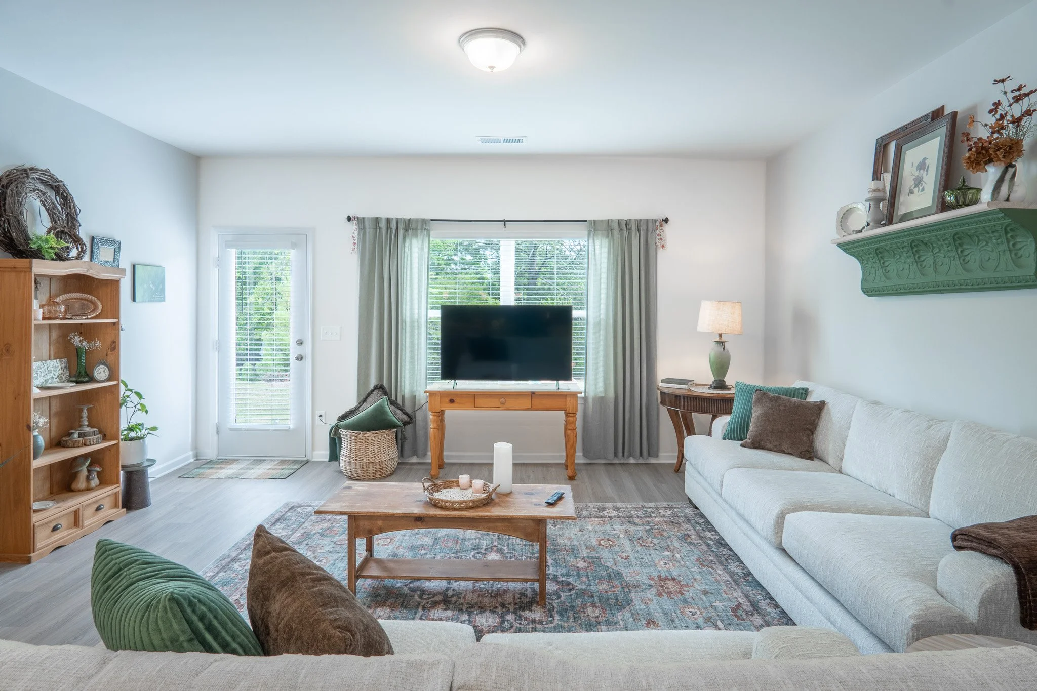 A living room with white walls, light wood flooring, a large white sofa with pillows, a wooden coffee table with candles, a flat-screen TV on a wooden stand, green and brown throw pillows, a patterned rug, a wooden bookshelf with decor, a door with b