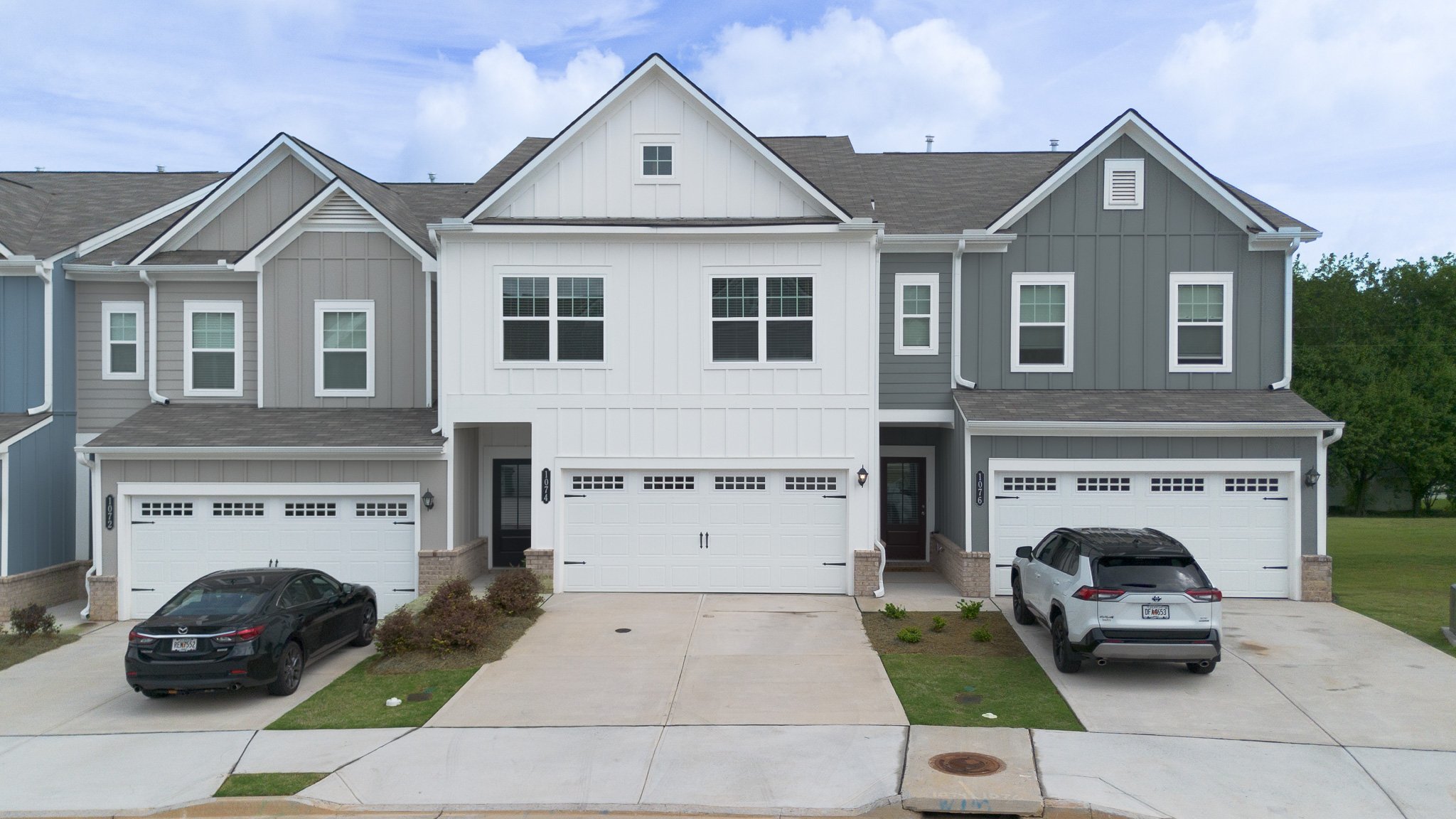 A row of modern townhouses with two car garages, three cars parked in the driveways, and multiple windows. The townhouses feature different siding colors: light blue, gray, and white. The sky is partly cloudy, and there are some small landscaping bus