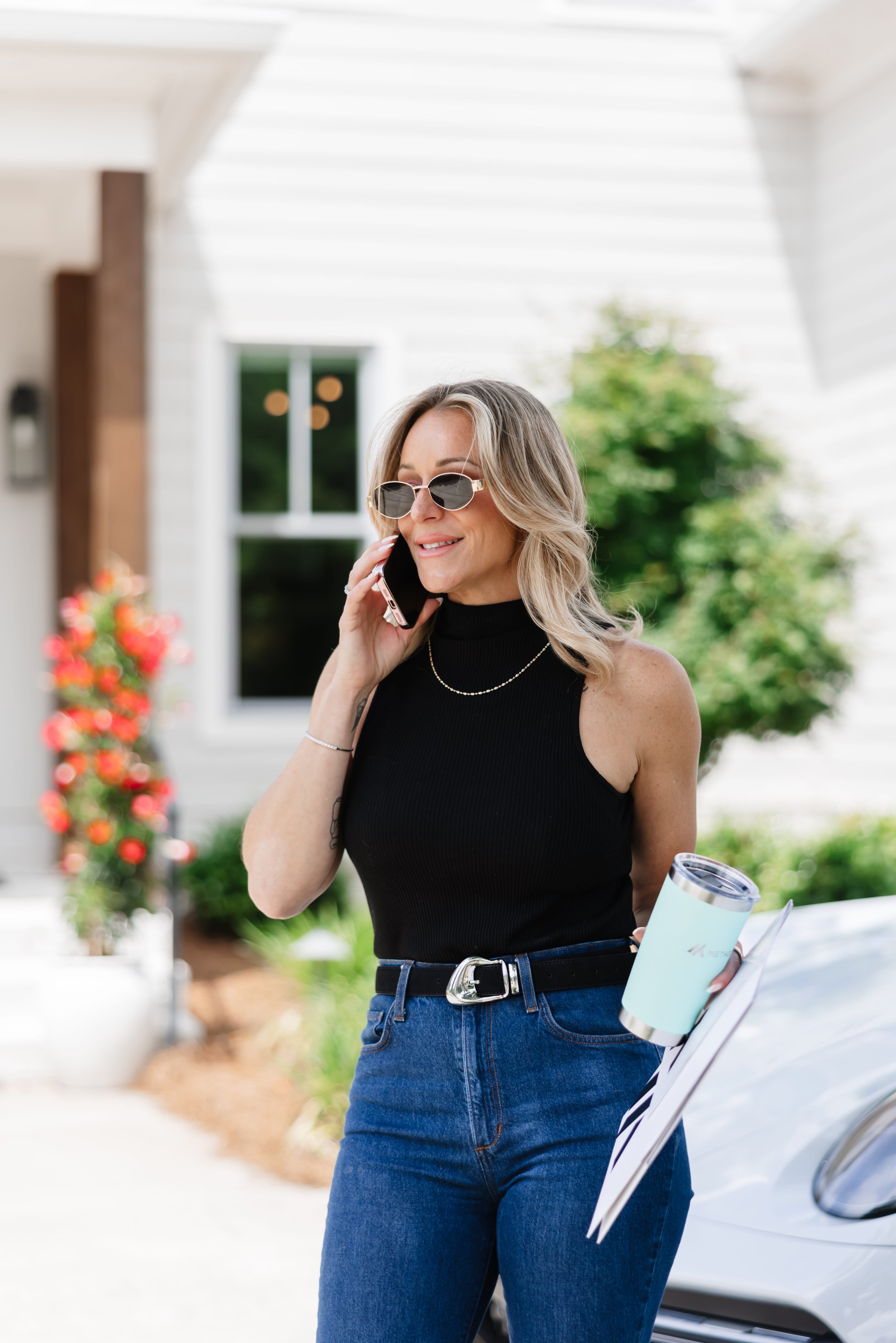 A woman with blonde hair, wearing sunglasses and a black sleeveless top, is talking on her phone while standing outdoors near a white car with a paper cup in her hand.