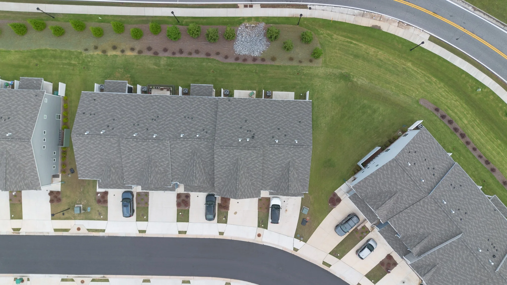 Aerial view of a residential street with neatly designed houses, a green lawn, and a park with trees and shrubs. Cars are parked along the street and in driveways, with a curved road and sidewalk visible.