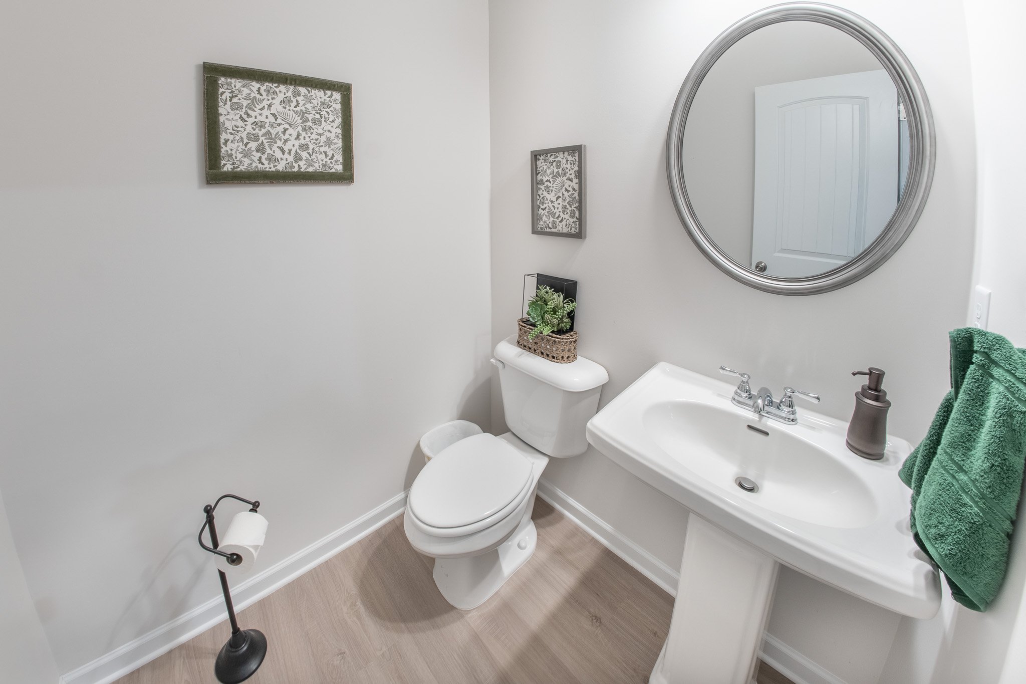 A small bathroom with a toilet, a pedestal sink with a mirror above, green towels, a small plant on the toilet tank, and two framed artwork pieces on the wall.
