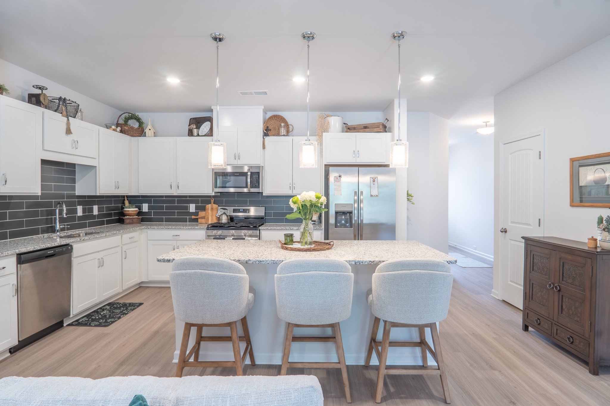 A modern kitchen with white cabinets, stainless steel appliances, a black backsplash, and a large island with a bouquet of white flowers. There are three white chairs at the island, pendant lights hanging above, and decorative items on top of the cab