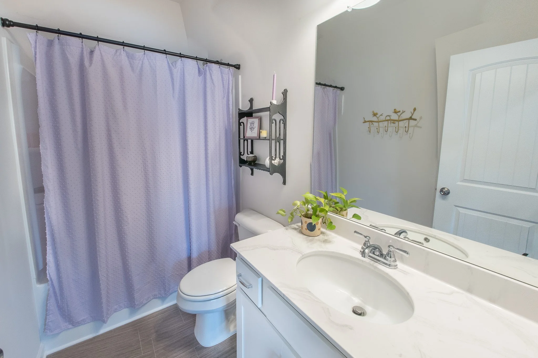A bathroom with a purple shower curtain, a white toilet, a white vanity with a sink, a large mirror, a black wall shelf with candles and framed art, a plant on the counter, and a gold wall hook with decorative hooks. The bathroom has beige walls and 