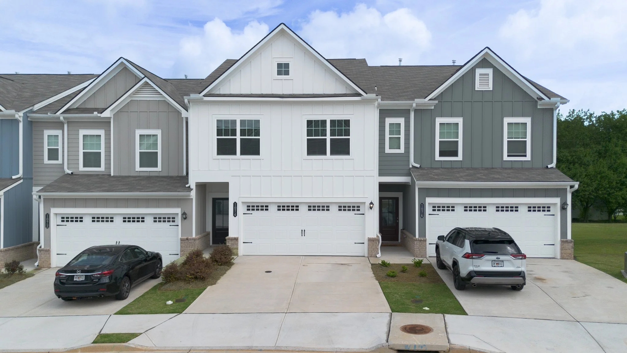 A modern three-story townhouse with two garages, gray and white siding, several windows, and a small front yard with parked cars.