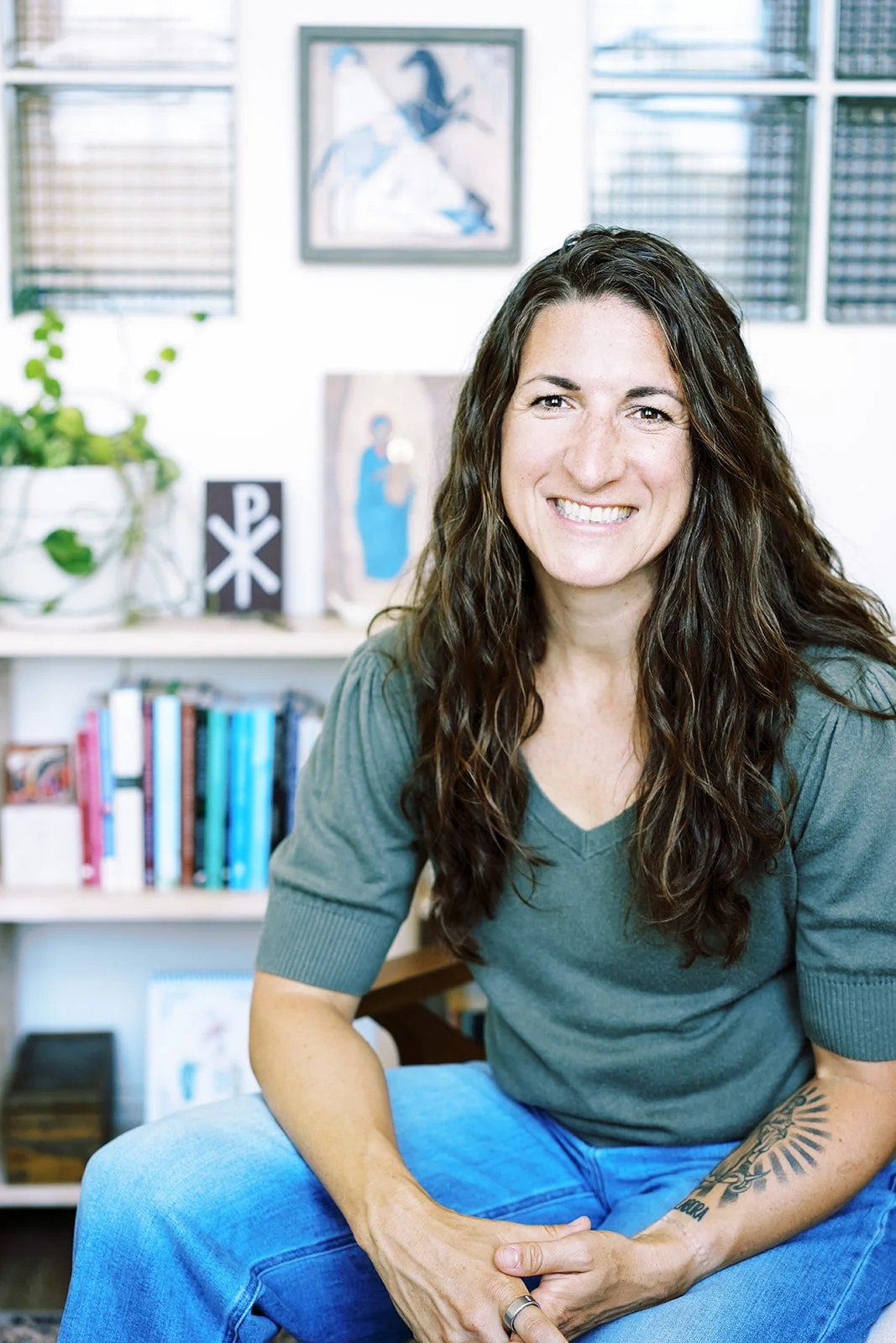 Kenda Mullert long curly dark hair and a tattoo on her forearm, smiling and sitting in a room with bookshelves and art on the wall.