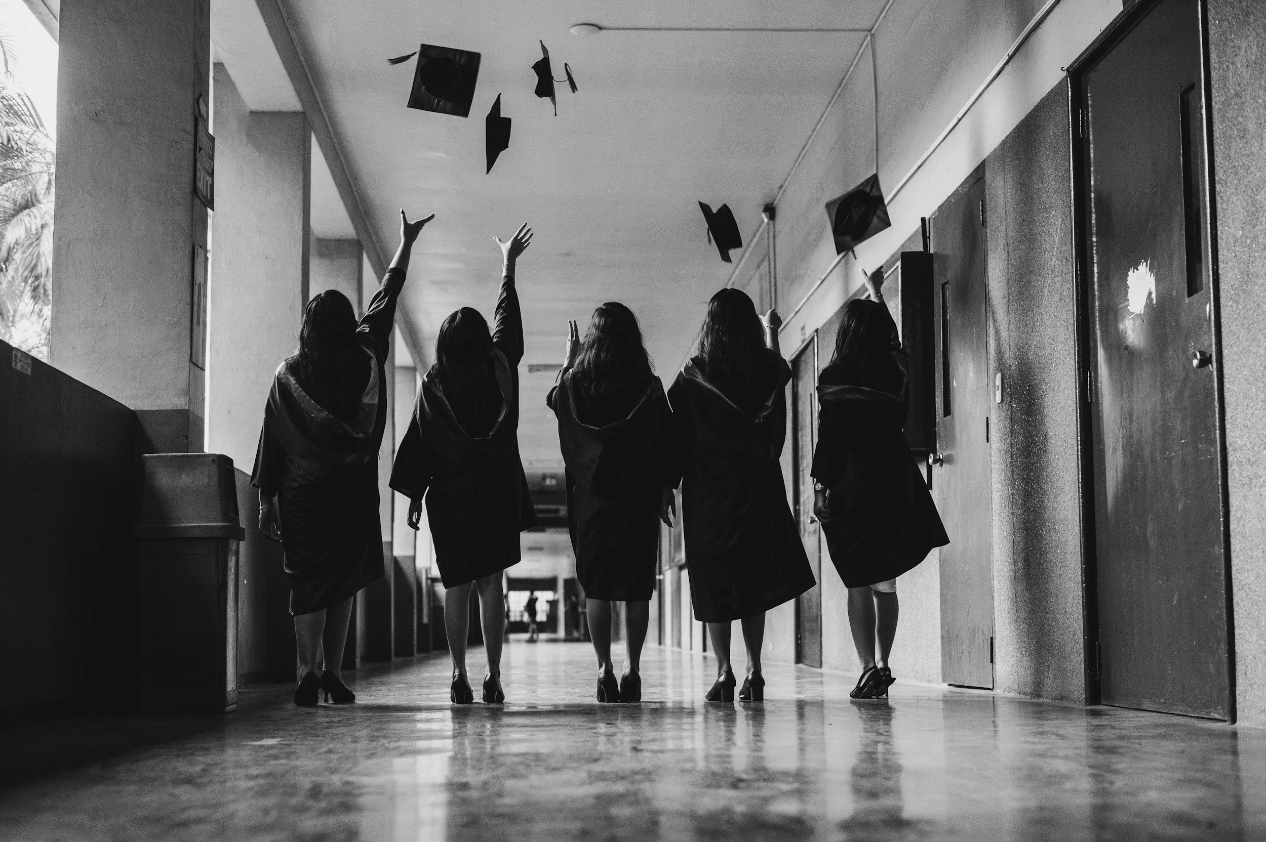 Four women in graduation gowns walking down a school hallway, throwing their graduation caps in the air.