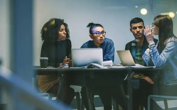 Four people in a meeting room engaged in a discussion, with laptops and notebooks on the table.