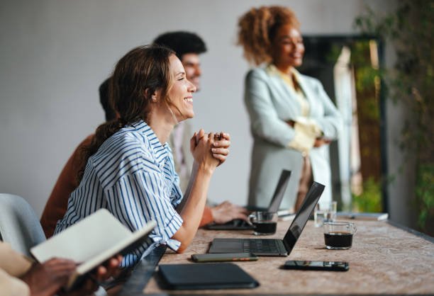Group of diverse women in a meeting room, smiling and engaging, with laptops and notebooks on the table.