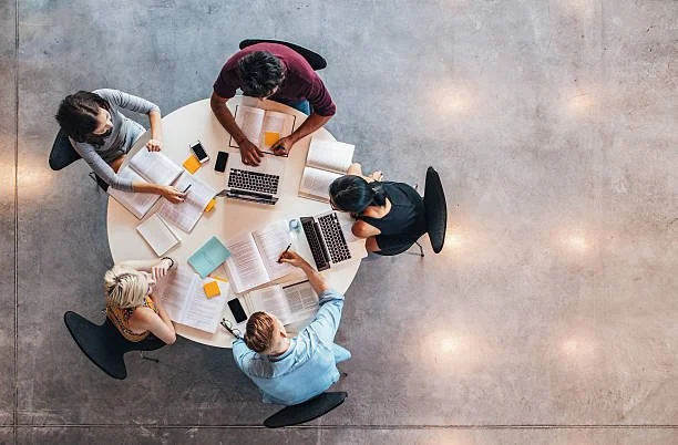 Top view of five people working together around a round table with laptops, notebooks, and books.