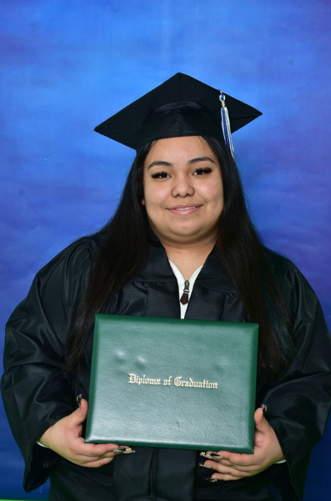 A young woman in a black graduation gown and cap holding a green diploma cover in front of a blue backdrop.