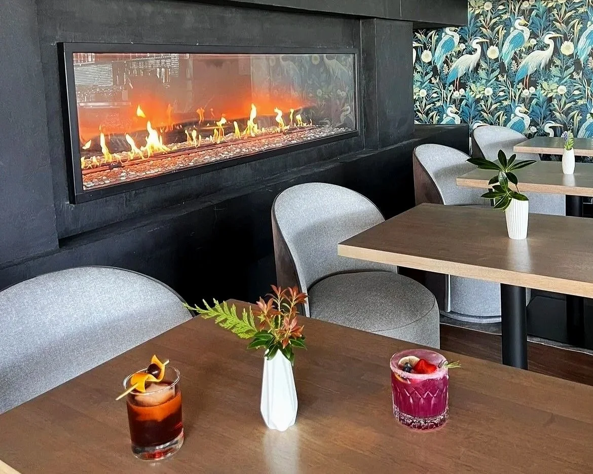 A restaurant interior with a modern fireplace, gray chairs, wooden tables, and small white vases with green plants. Commercial Design from Spirit design studio - Vancouver island interior decorator